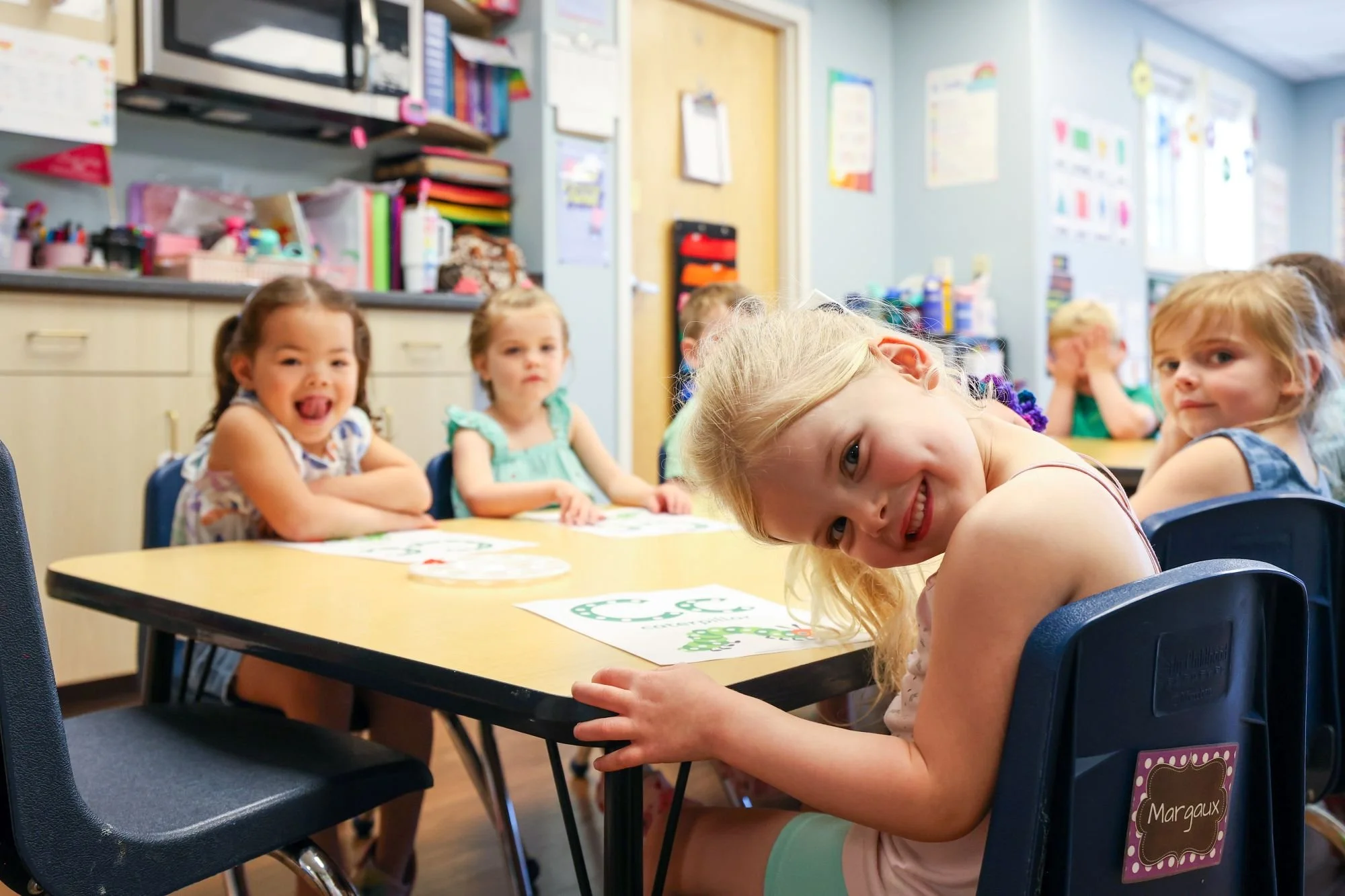 Young children sitting at a table in a classroom, smiling and looking at the camera. The classroom has colorful educational posters and supplies in the background.