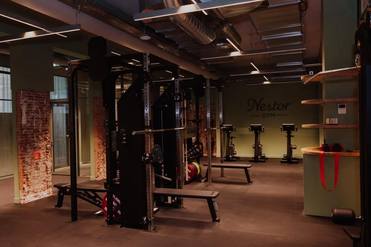 Innenraum eines Fitnessstudios mit Kraft- und Cardiogeräten, die Wände sind teilweise aus Backstein, die Wände sind grün und an der Wand steht 'Nestor GYM'.