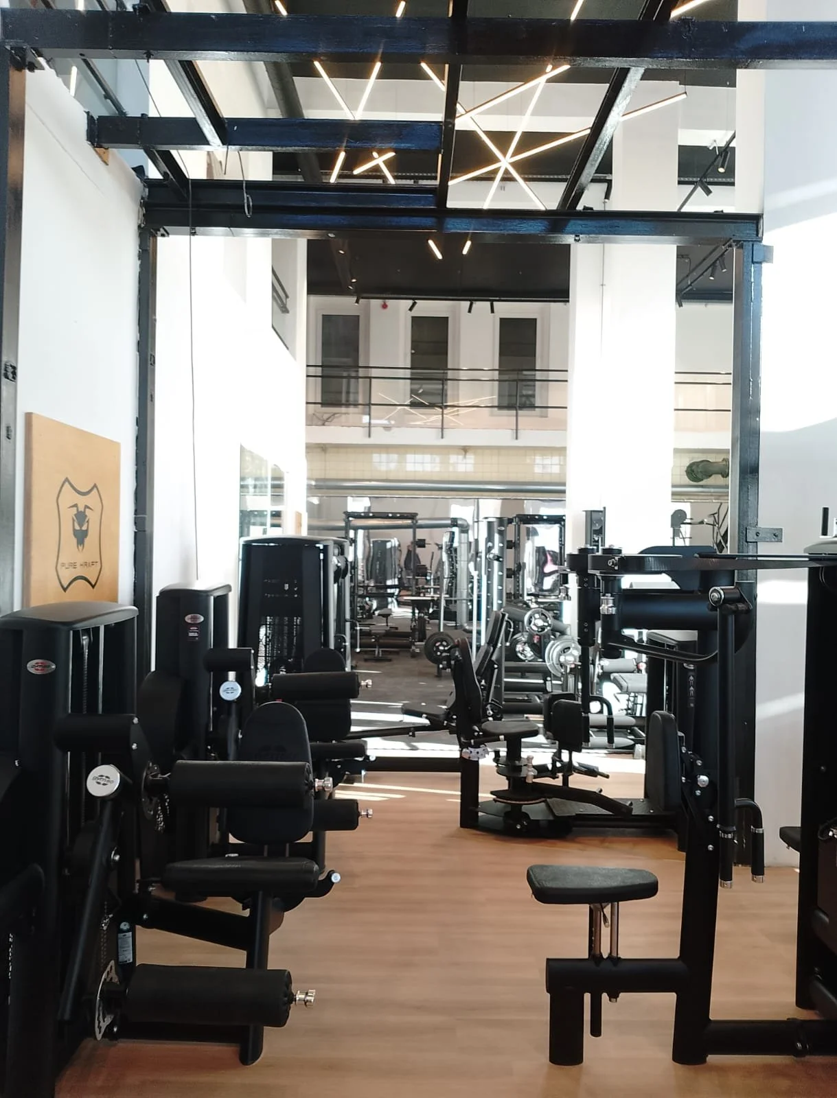 Innenraum eines Fitnessstudios mit verschiedenen Kraft- und Ausdauergeräten, Holzboden, weißen Wänden und einem modernen Beleuchtungsdesign an der Decke.
