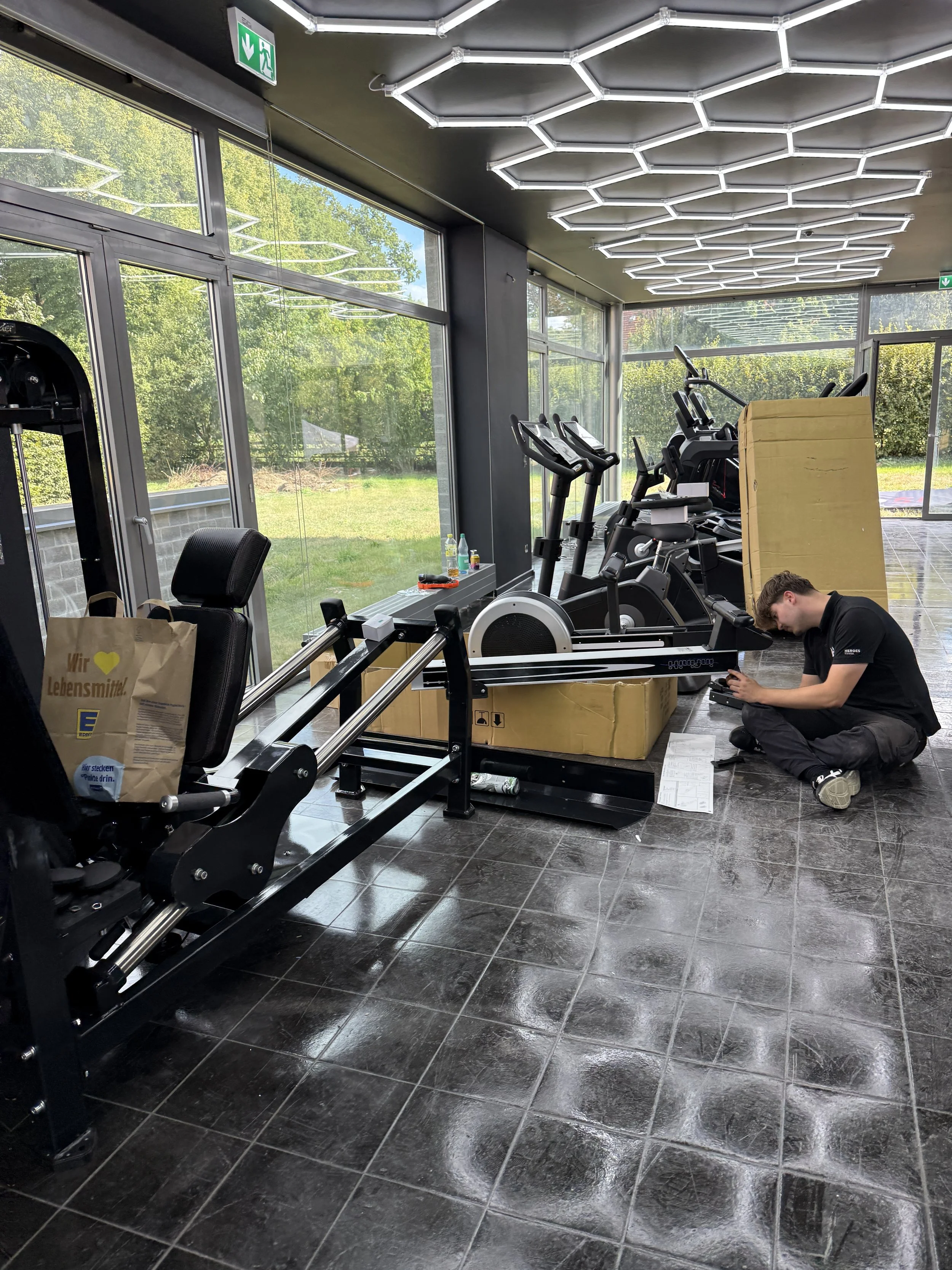 Fitnessraum mit verschiedenen Trainingsgeräten, während dein Techniker am Boden sitzt und ein Gerät montiert. Im Hintergrund stehen weitere Cardio- und Kraftgeräte.