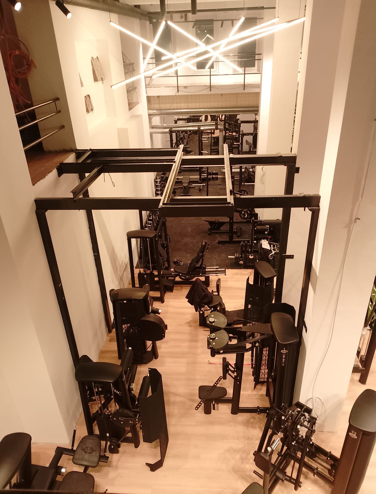 Ein schmaler Fitnessraum mit verschiedenen Trainingsgeräten, darunter ein stationäres Fahrrad, eine Sitzmaschine, Gewichte und Hanteln, mit Holzfußboden und moderner Beleuchtung.
