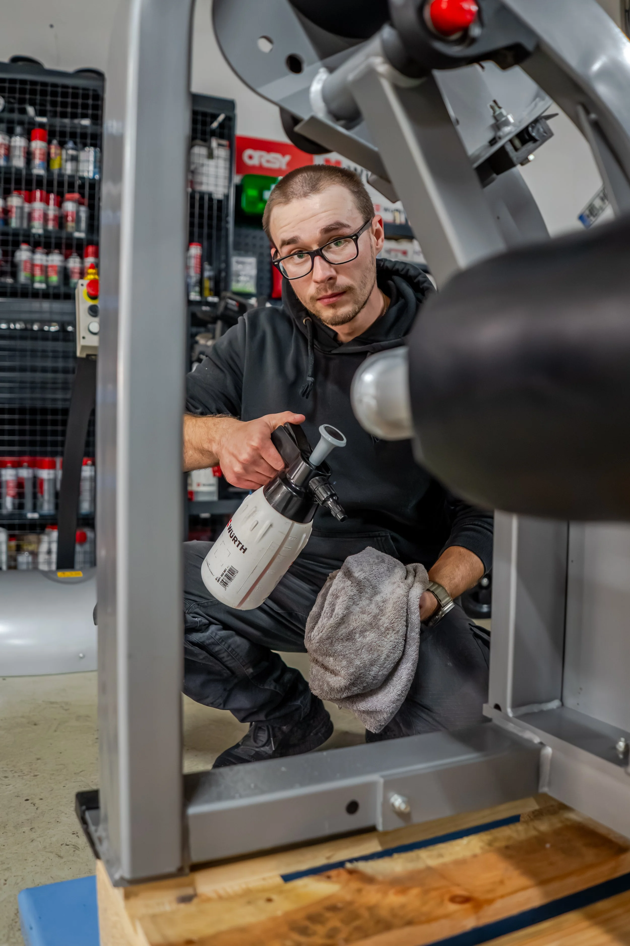 Ein Mann mit Brille und dunkler Kleidung inspiziert eine Maschine in einem Werkzeugladen oder einer Werkstatt, während er ein Sprayflasche hält und eine graue Tücher in der Hand hat.