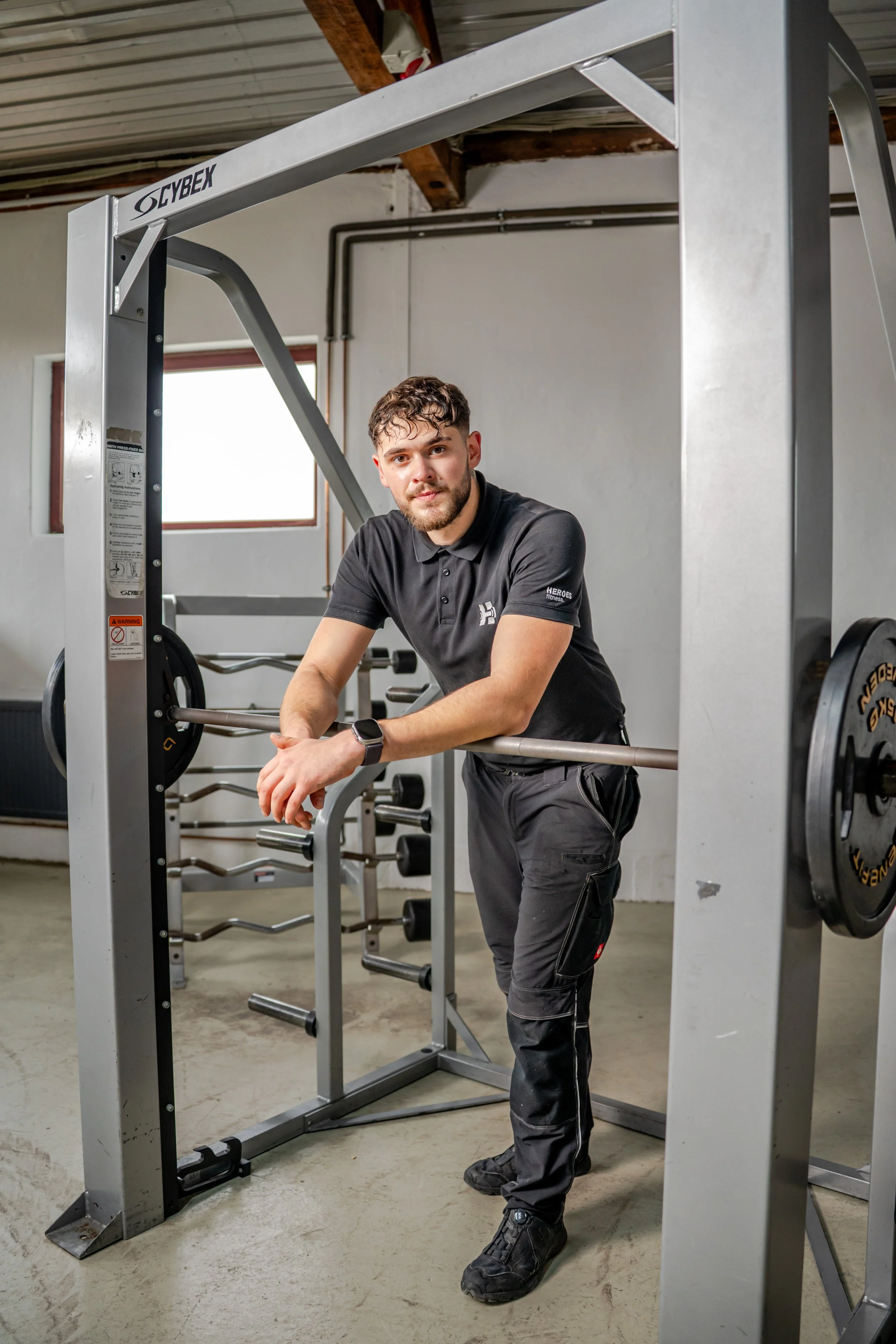 Junger Mann steht in einem Fitnessstudio und lehnt sich auf eine Hantelstange, umgeben von Trainingsgeräten.