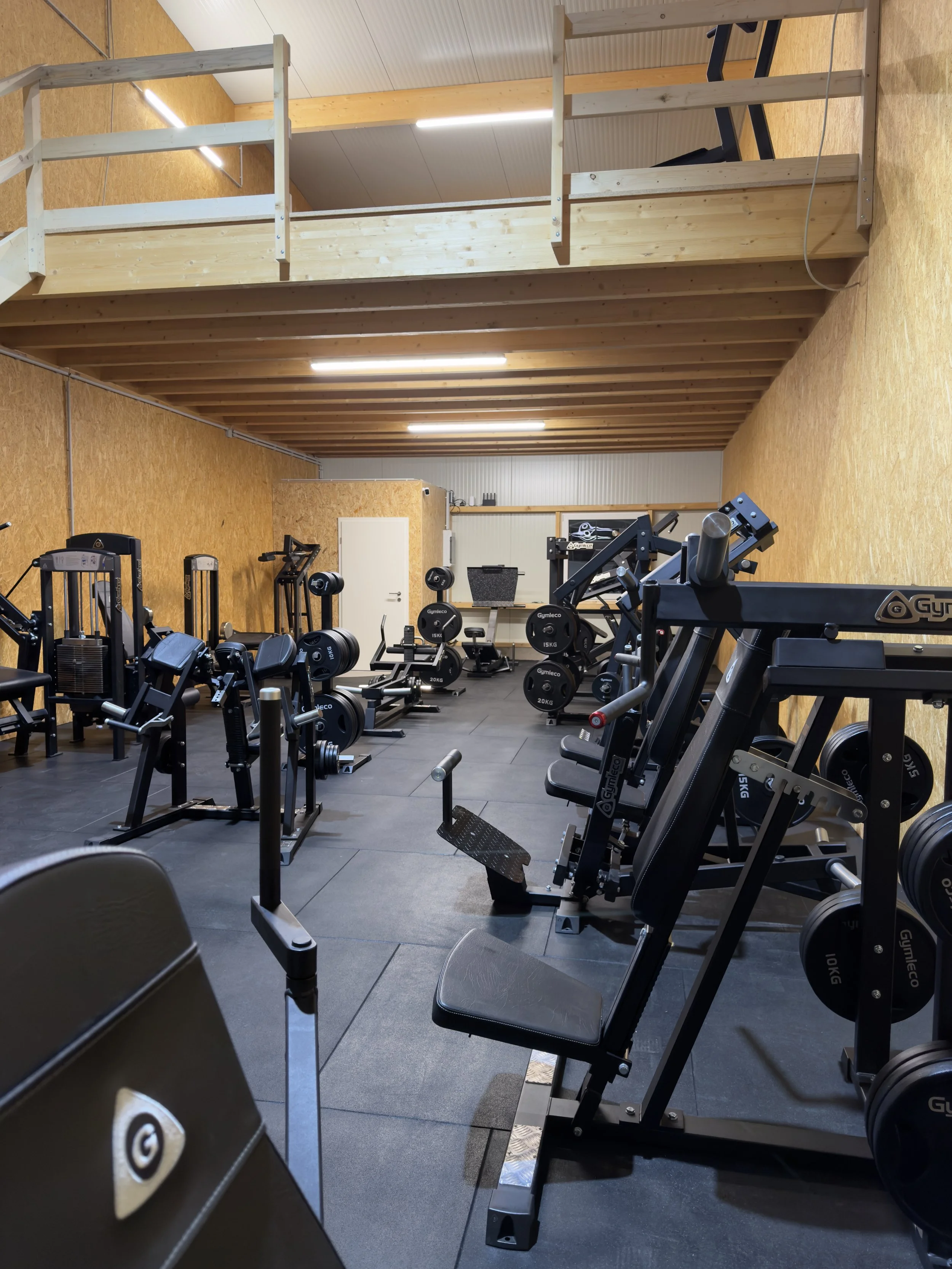 Ein Fitnessstudio mit verschiedenen Krafttrainingsgeräten, Holzwänden und einer oberen Empore aus Holz.