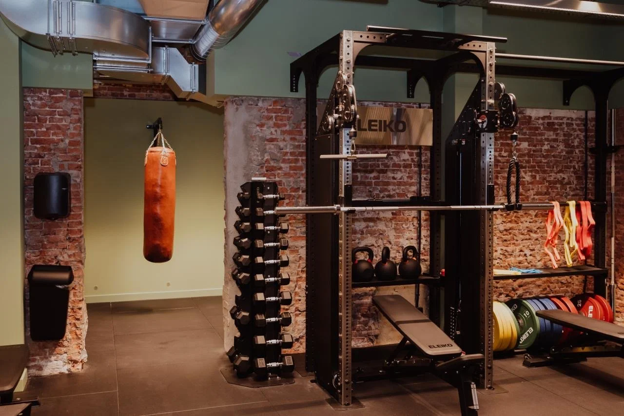 Fitnessraum mit Hantelbank, Kurzhanteln, Gewichtsscheiben, Kettlebells, Langhantel, Boxsack, und Widerstandsbänder vor einer Ziegelwand.