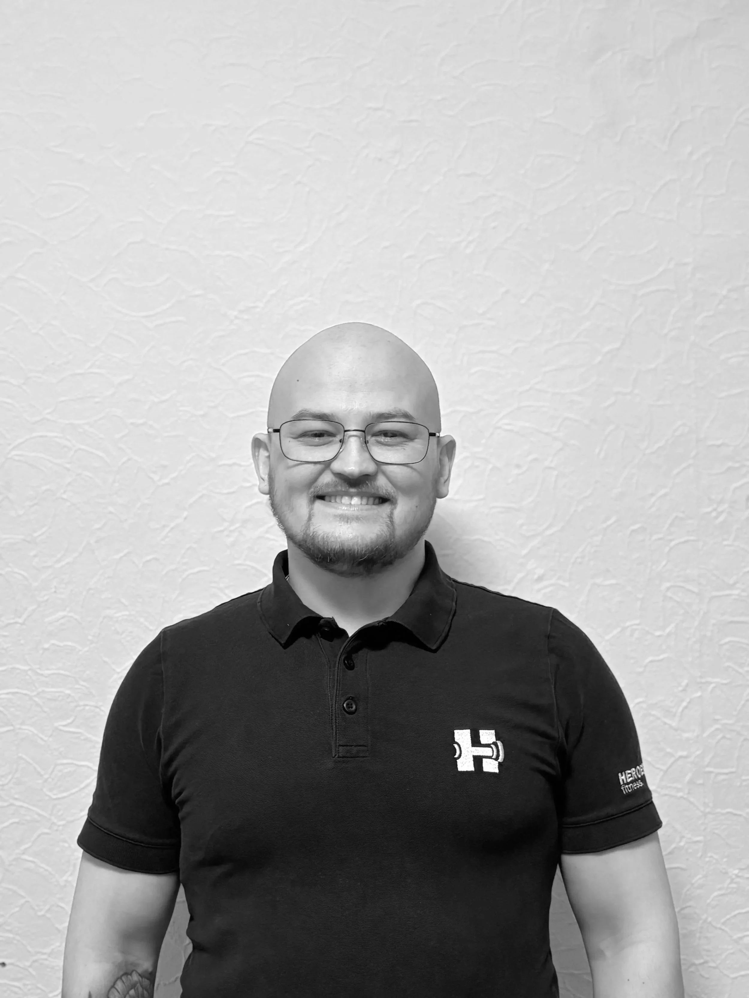Ein lächelnder Mann mit Brille, Bart und Glatze trägt ein schwarzes Polo-Shirt mit Logos auf der Brust.