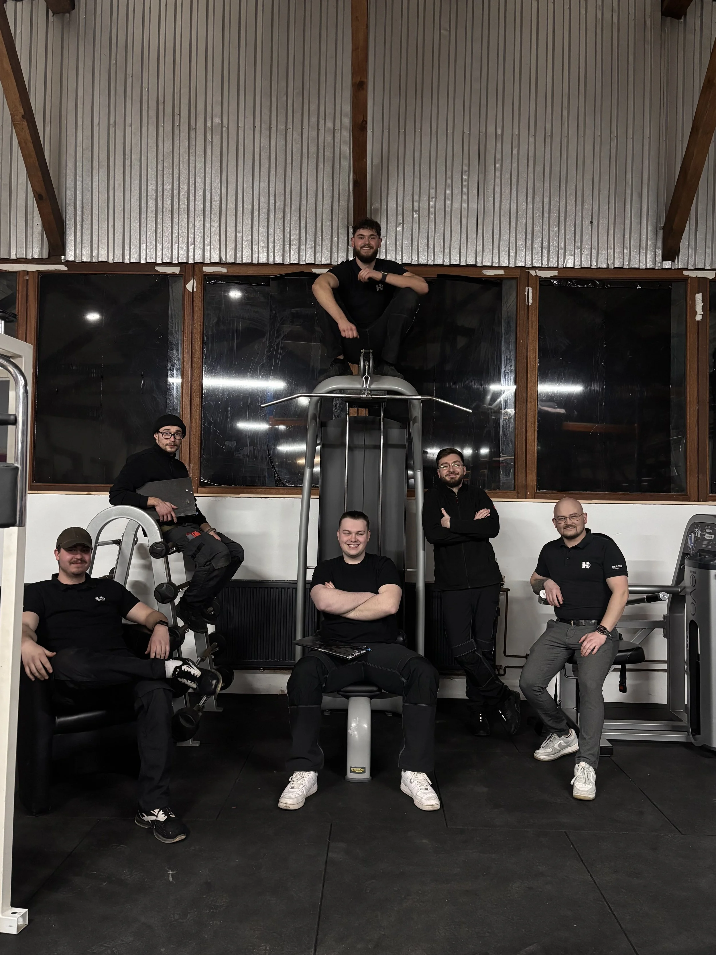 Sechs Männer posieren in einem Fitnessstudio, einer sitzt auf einem Trainingsgerät, einer auf einer Leiter, einer auf einem Stuhl, eine Person steht mit verschränkten Armen, eine steht mit einem Laptop, und einer sitzt auf einem Bank.