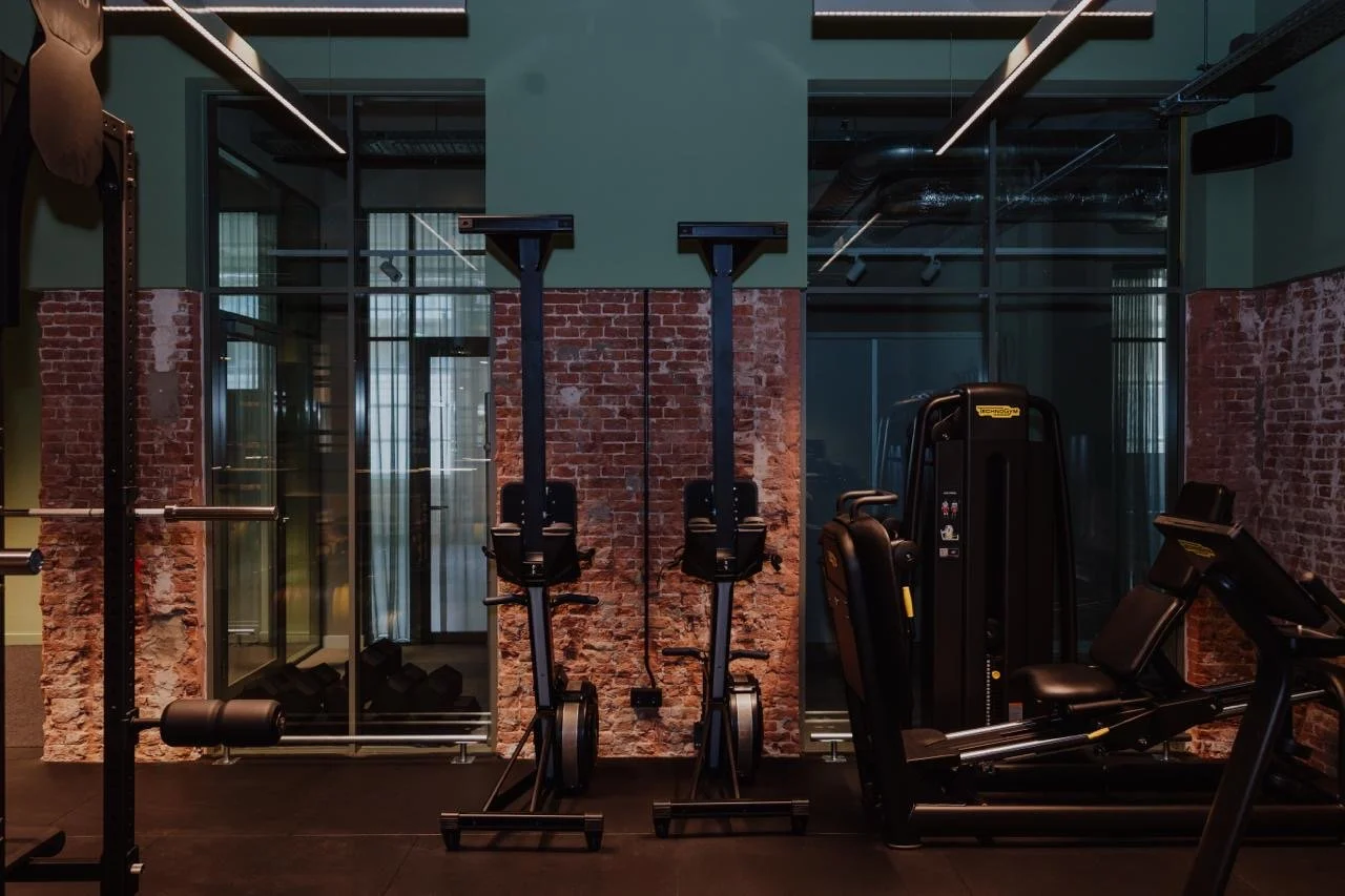 Fitnessstudio mit verschiedenen Gym-Ausrüstungen wie Laufbändern, Rudergeräten und Krafttrainingsgeräten, vor Ziegelwand und Fenstern mit Vorhängen.