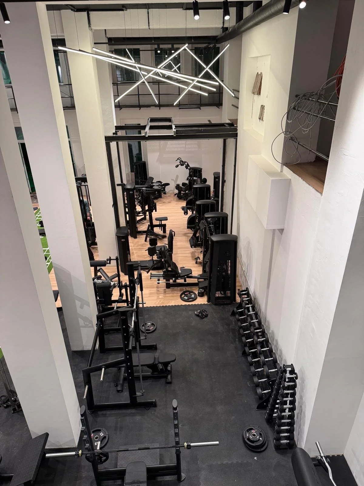 Fitnessstudio mit verschiedenen Geräten und Hanteln, modernes Design mit weißen Wänden und schwarzen Trainingsgeräten, Deckenbeleuchtung und Holztechnik im Hintergrund.