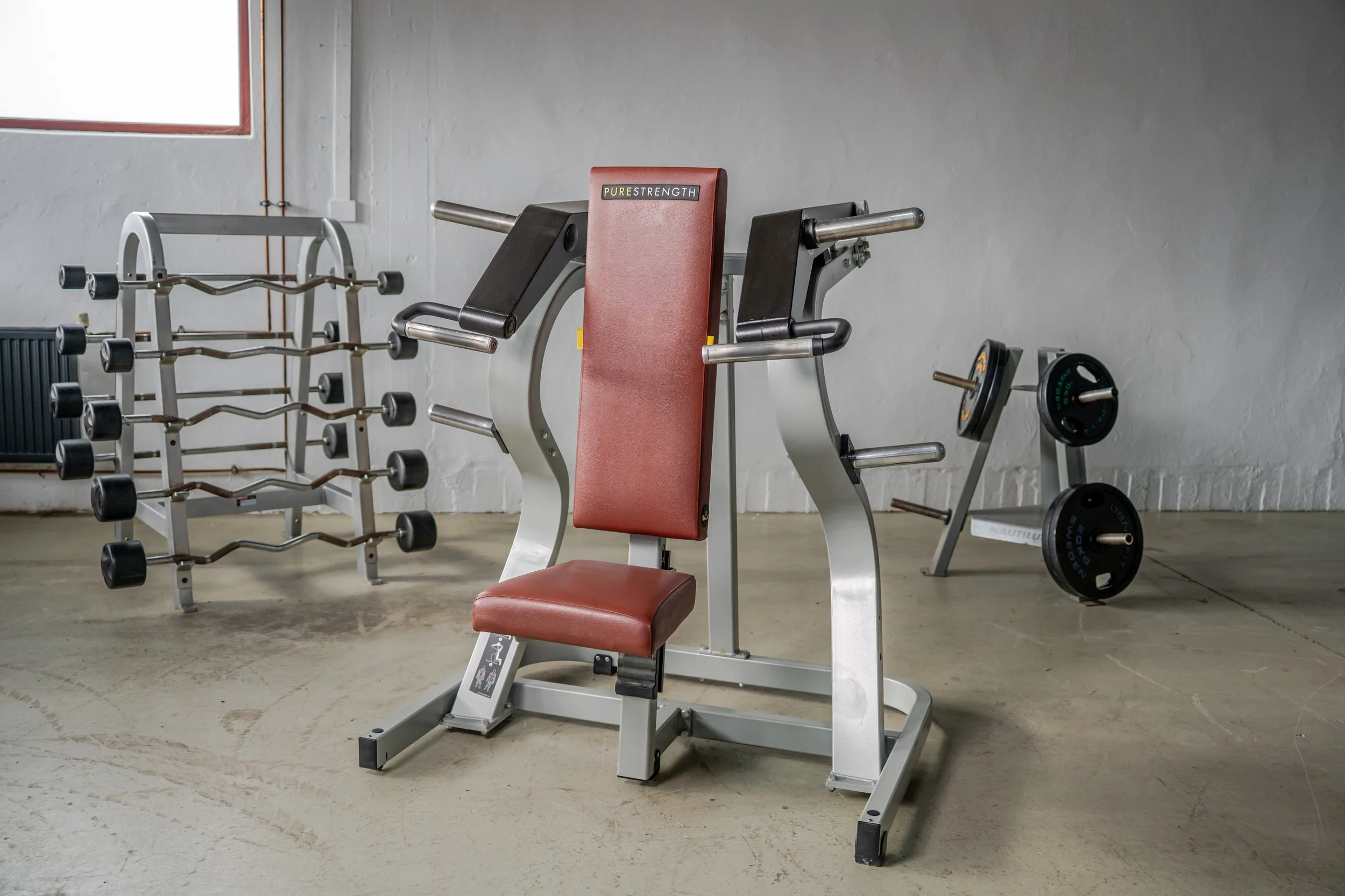 Fitnessgeräte in einem Fitnessstudio, darunter eine sitzende Rückenstreckermaschine, Hanteln auf einem Racks, und eine Langhantel auf einem Ständer, in einem Raum mit weißen Wänden und einem Fenster.