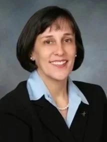 A professional woman with short dark hair wearing a black blazer, light blue dress shirt, and pearl necklace, smiling against a gray background.