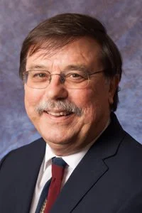 Portrait of a middle-aged man with brown hair, glasses, and a mustache, wearing a dark suit, white shirt, and a patterned tie, against a blue background.