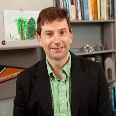 A man with short brown hair wearing a black blazer and light green shirt, standing in front of bookshelves and a drawing of a palm tree.