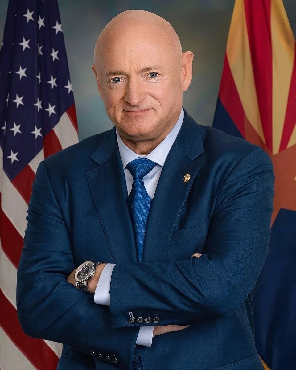 A bald man in a dark blue suit, white shirt, and blue tie standing with arms crossed, in front of American and Arizona flags.