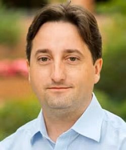 Headshot of a man with dark hair, wearing a light blue collared shirt, outdoors in a park setting.