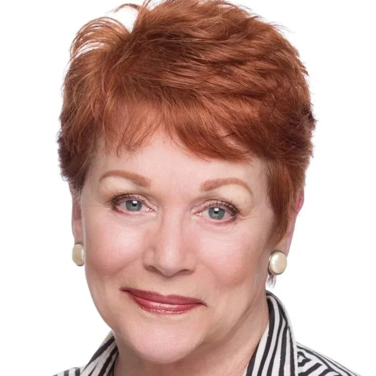 Close-up portrait of a middle-aged woman with short red hair, wearing pearl earrings, and a black and white striped shirt against a white background.