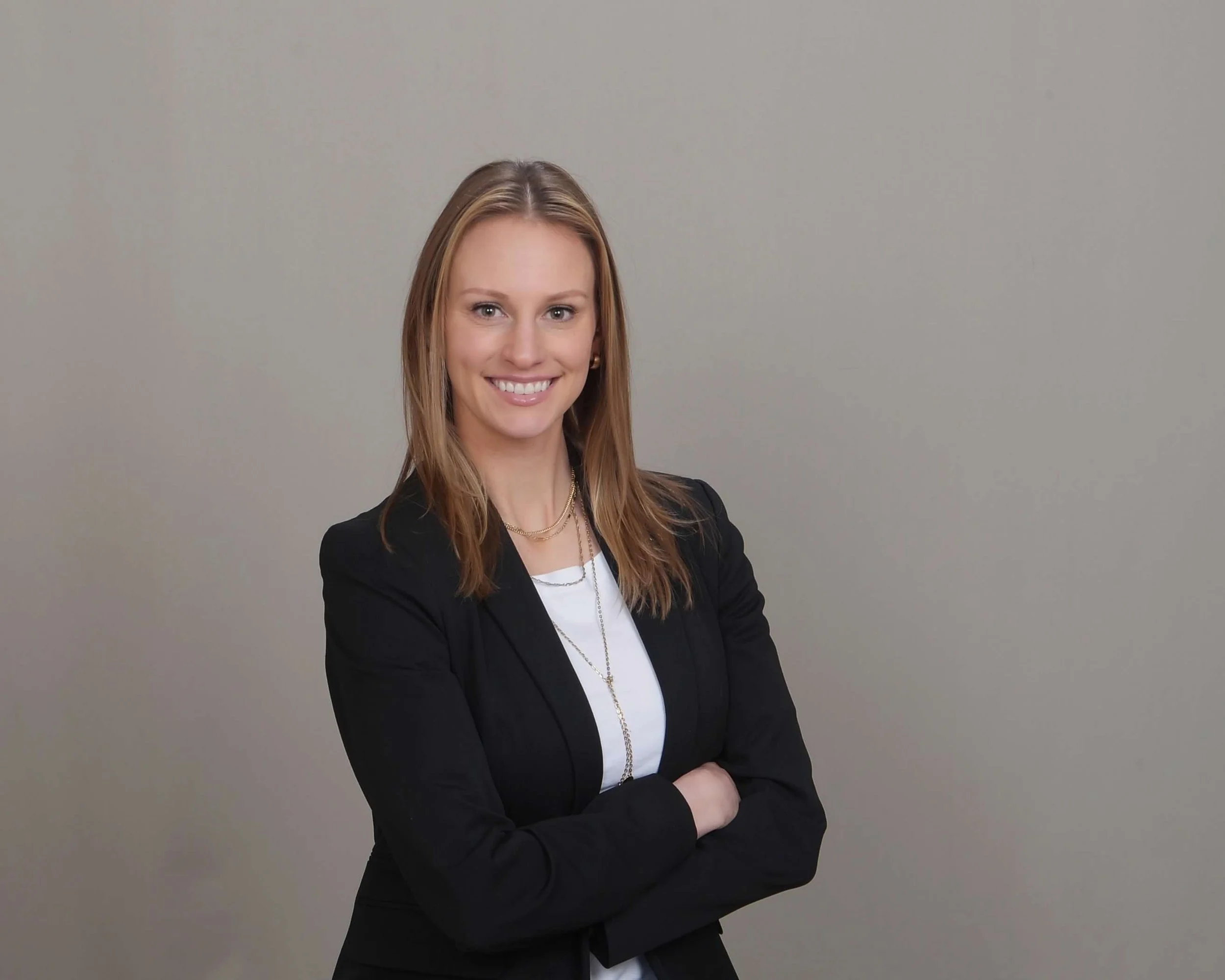 A woman with shoulder-length light brown hair, wearing a black blazer over a white top, standing with arms crossed and smiling against a plain, light-colored background.