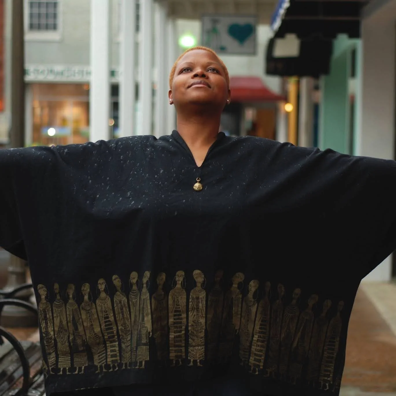 A person with short hair raising their arms in a confident pose, wearing a black garment with gold tribal figures at the bottom, standing in an indoor shopping or dining area.