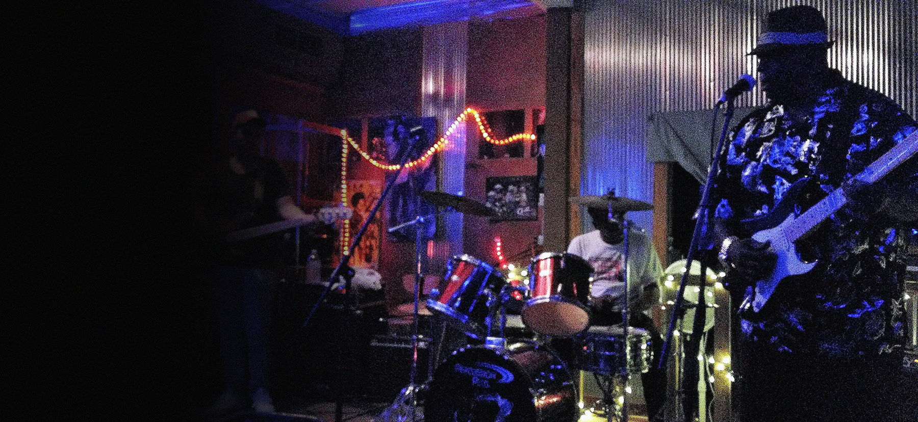 A band performing live music at an indoor venue with dim lighting, neon string lights, and acoustic wall panels. The band includes a keyboard player, drummer, and guitarist, with the guitarist wearing a hat and sunglasses.
