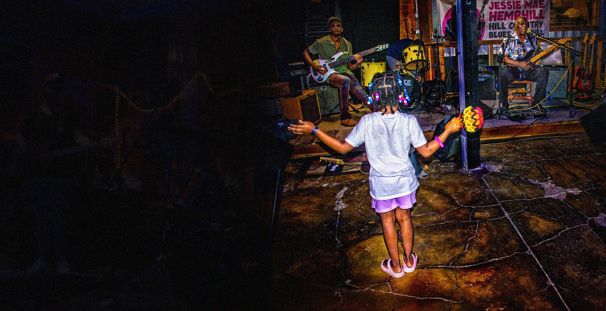 A young girl with braided hair and beads, wearing a white t-shirt and pink shorts, dancing in front of a stage with live musicians playing guitars.