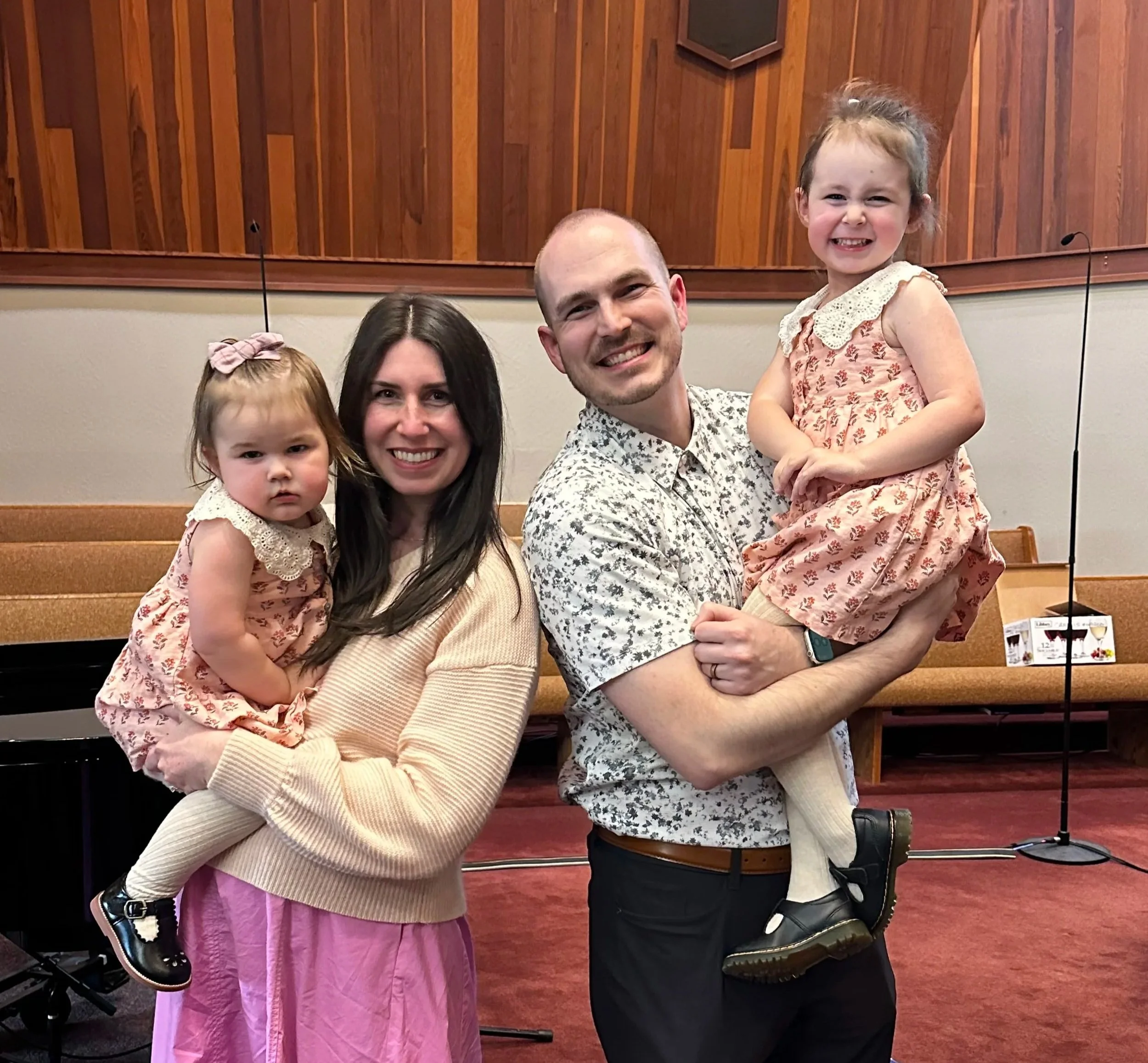 Four people, one being Pastor Zak Graves, two adults and two young girls, posing inside a church or chapel with wooden walls and theatre-style seating in the background. The man and woman are each holding a girl. All are smiling or looking content.