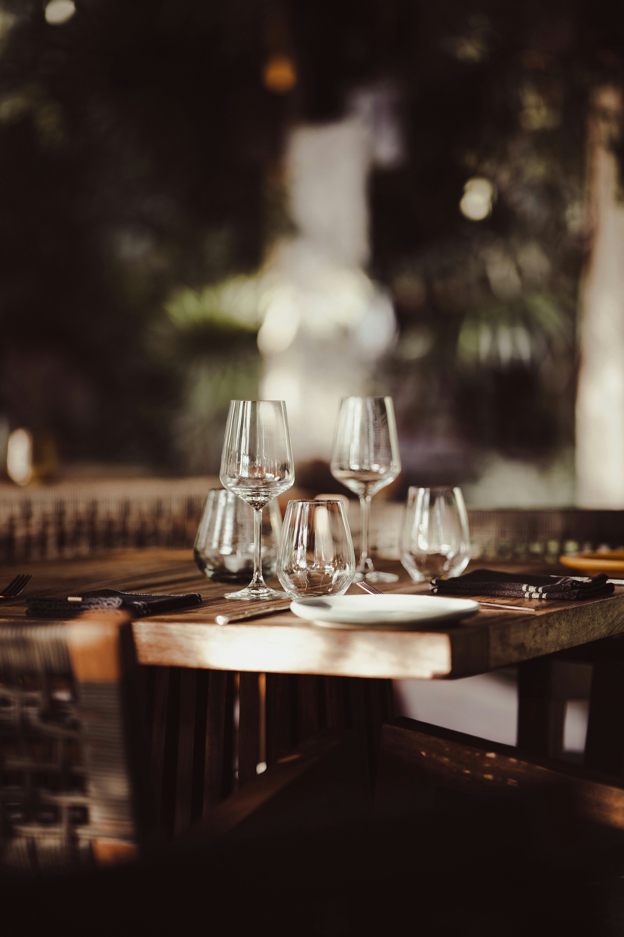 Empty wine glasses and water glasses placed on a wooden dining table with a white plate, black napkins, and silverware, set for a meal at an outdoor restaurant or patio.