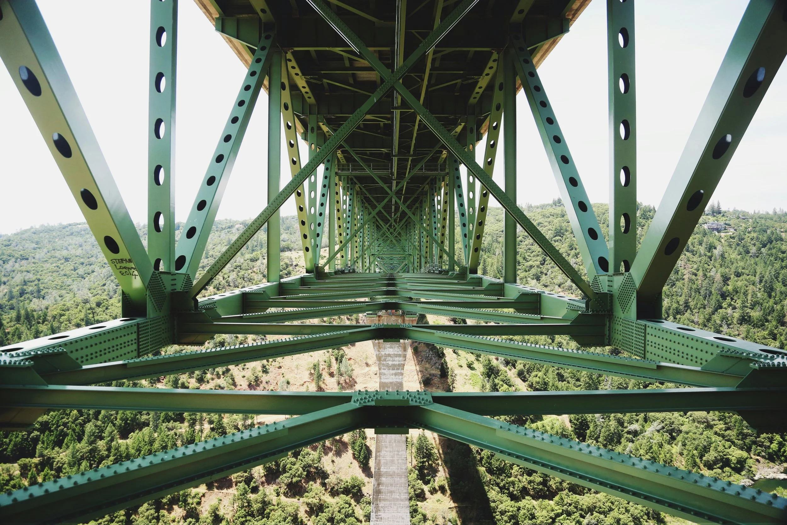 Looking up from beneath a green steel bridge, showing its intricate framework and support beams, with a lush, green landscape and a distant river below.