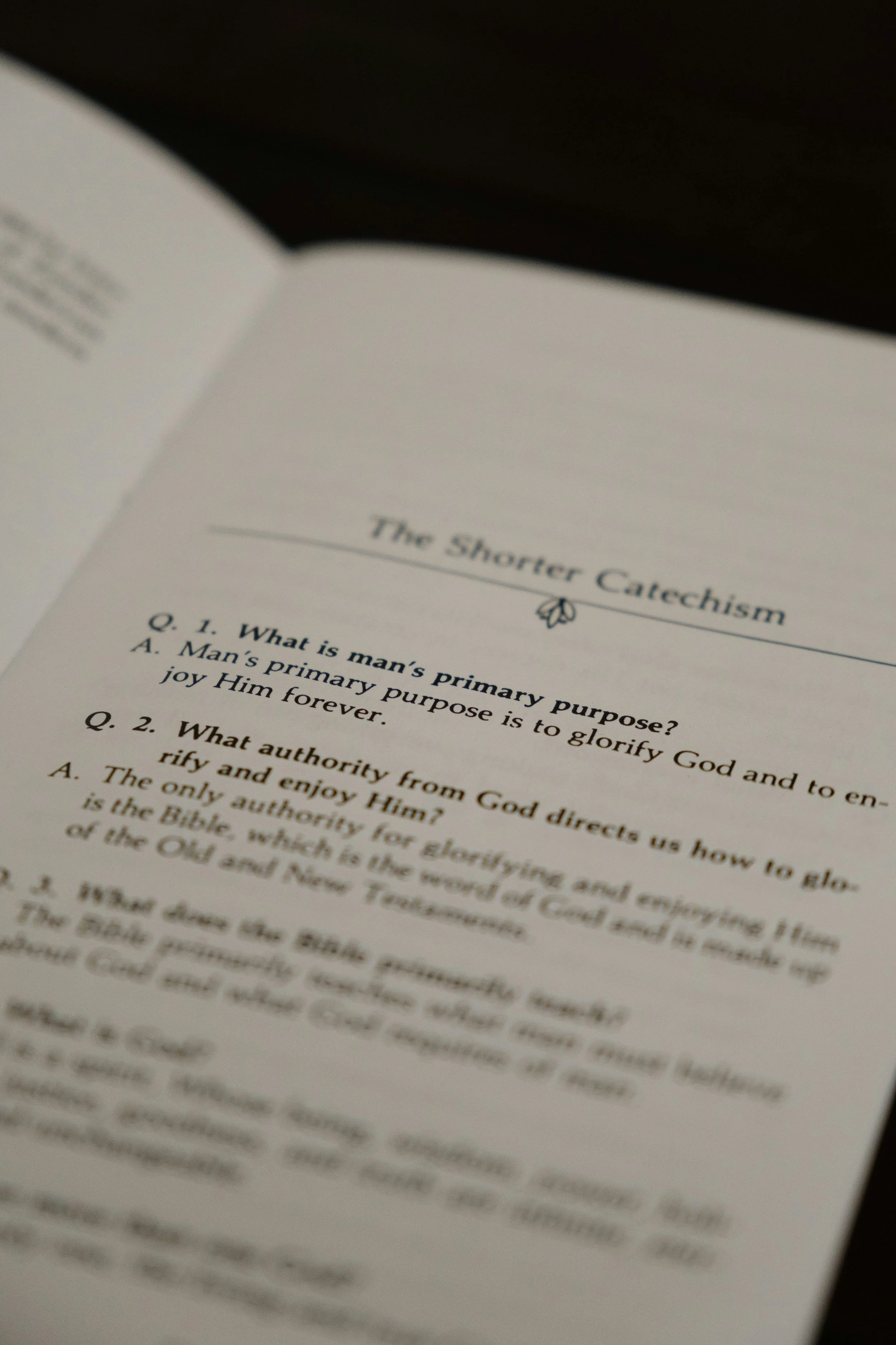 An open book page with the title 'The Shorter Catechism' and questions about God's purpose and authority, with a small decorative emblem above the title.