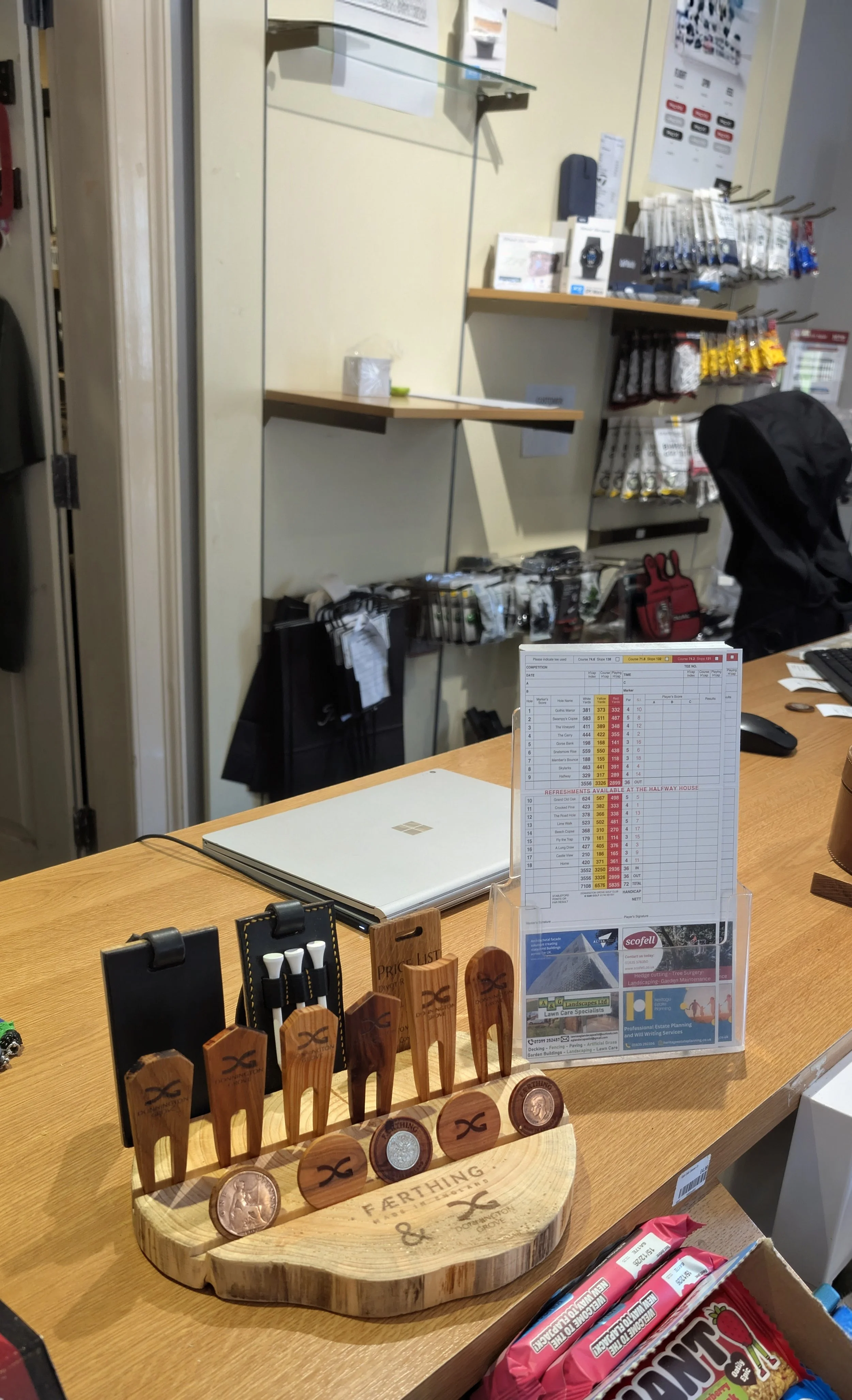 A wooden display stand with golf tees and coins on a store counter, with various office supplies, price list, and retail items in the background. In the Donnington Grove Golf Club pro shop.