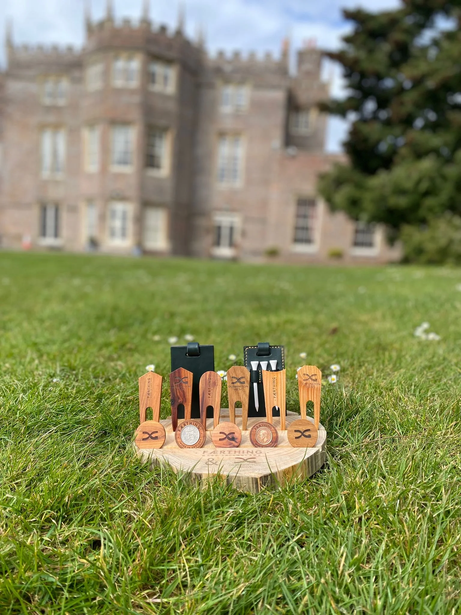 A set of small wooden golf tees with a leather case, placed on a wooden slab, on a grassy lawn with a blurred castle-like building in the background.