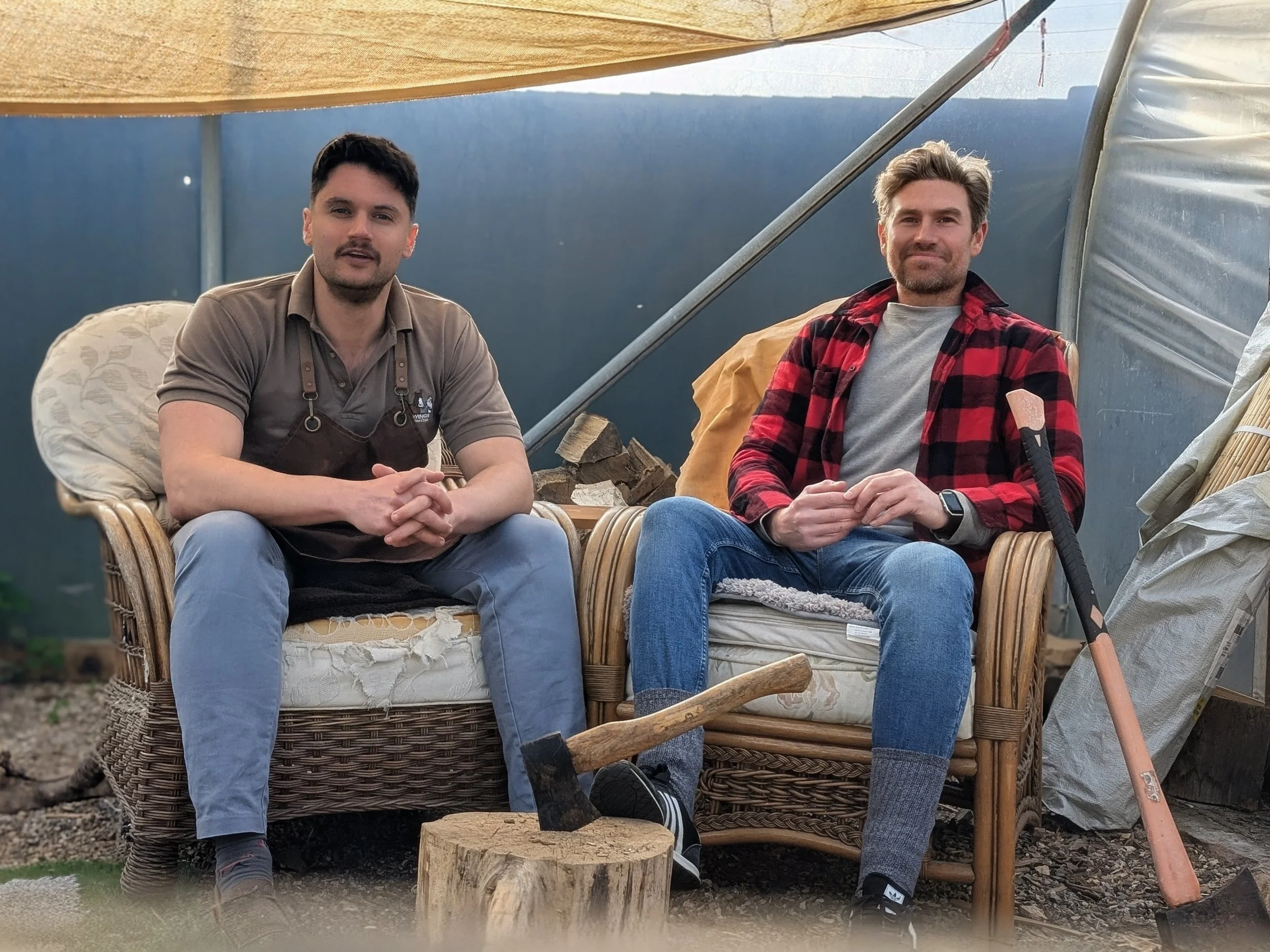 Two men sitting in wicker chairs inside a tent or greenhouse, surrounded by logs and gardening tools.