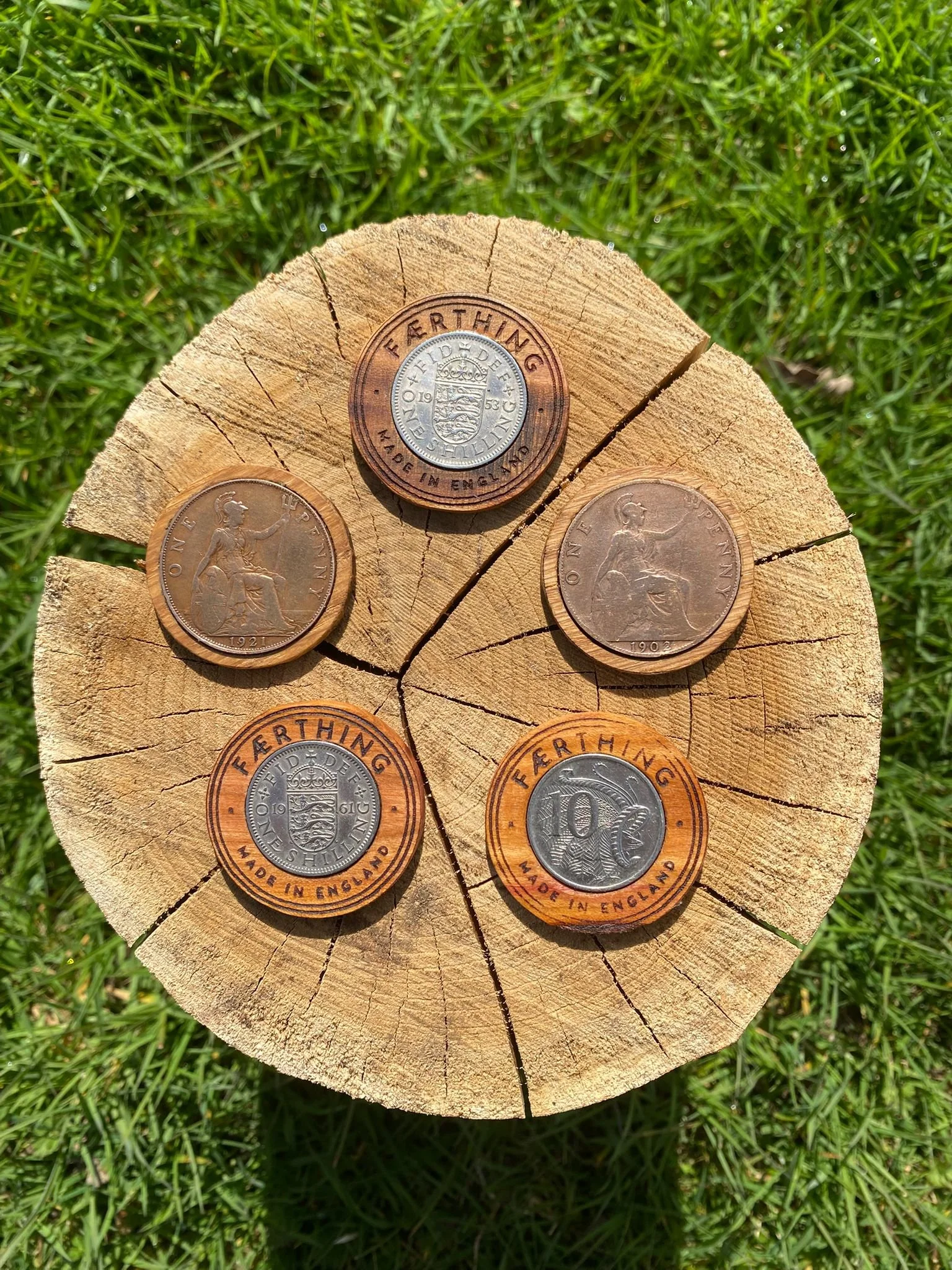 A wooden disk with five UK coins attached, including two one-penny coins, two five-pence coins, and one ten-pence coin, all labeled with the word "Fertinging".