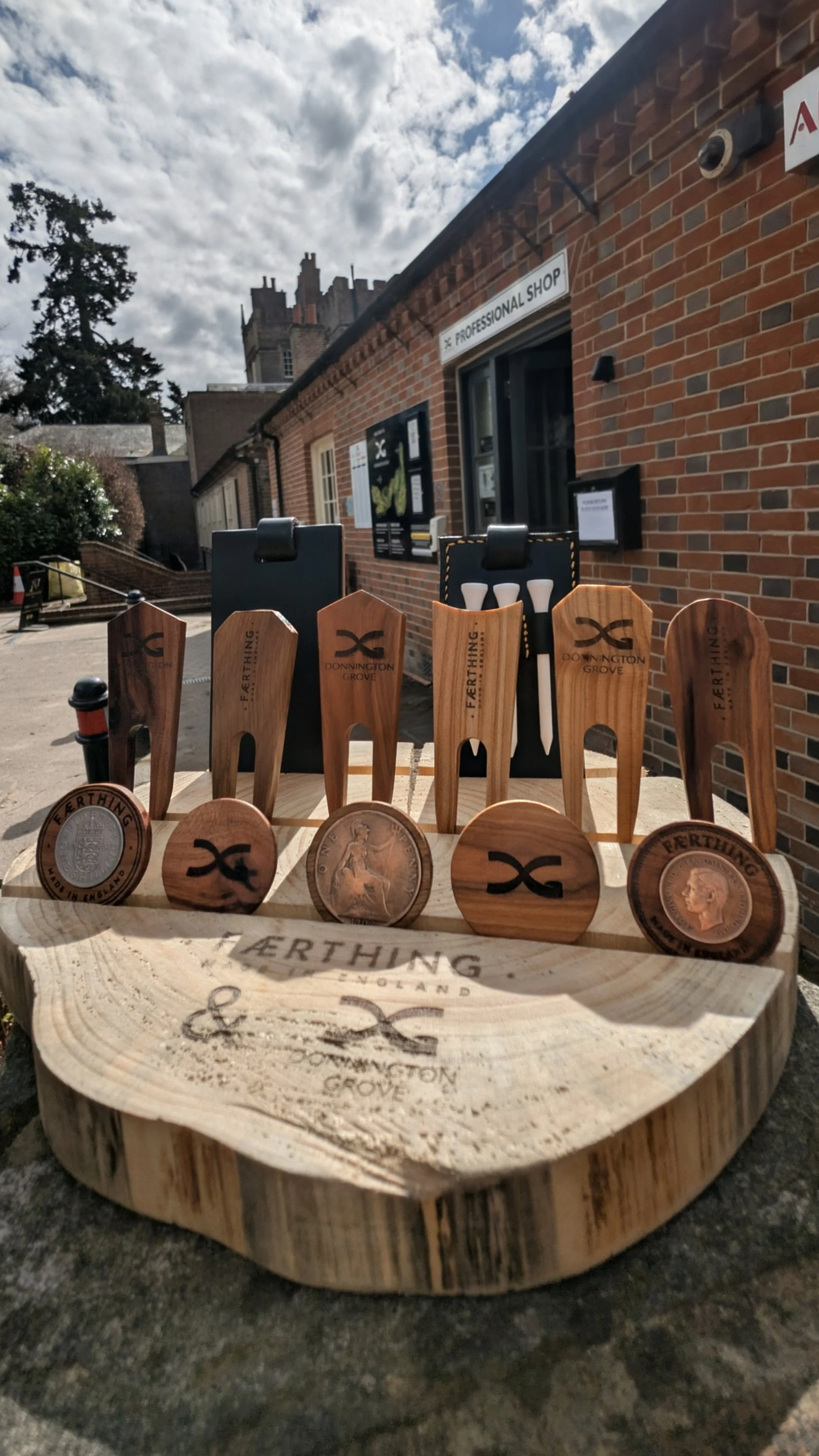 A display of wooden planting tools, such as shovels and trowels, with some bearing the 'Donnington Grove' logo, arranged on a carved wooden slab outside a brick building labeled 'Professional Shop' under a partly cloudy sky.