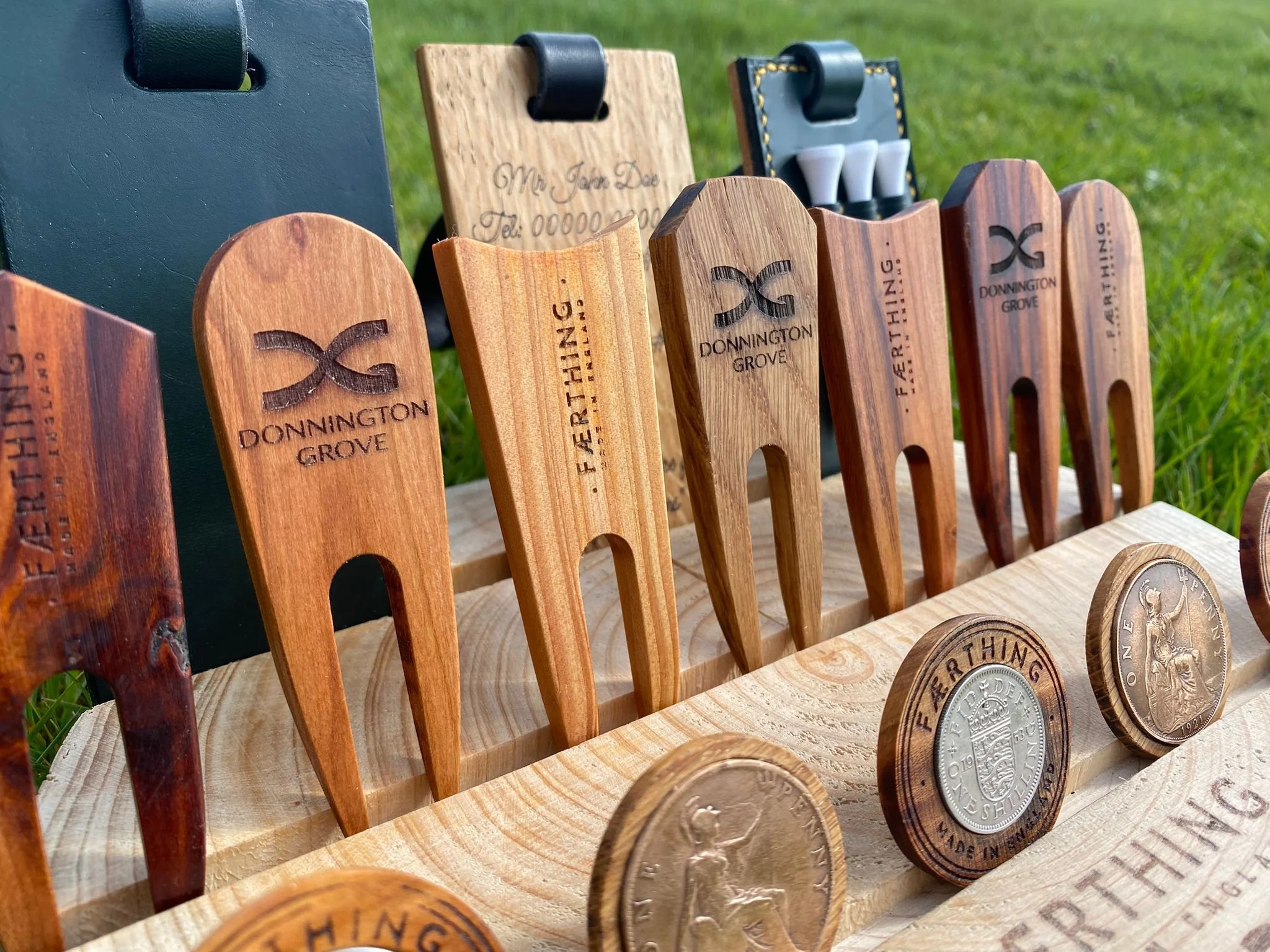 A display of wooden and metal golf accessories, including wooden golf tees engraved with 'Donnington Grove' and a metal coin with a seated figure, on a wooden stand outdoors on grass.