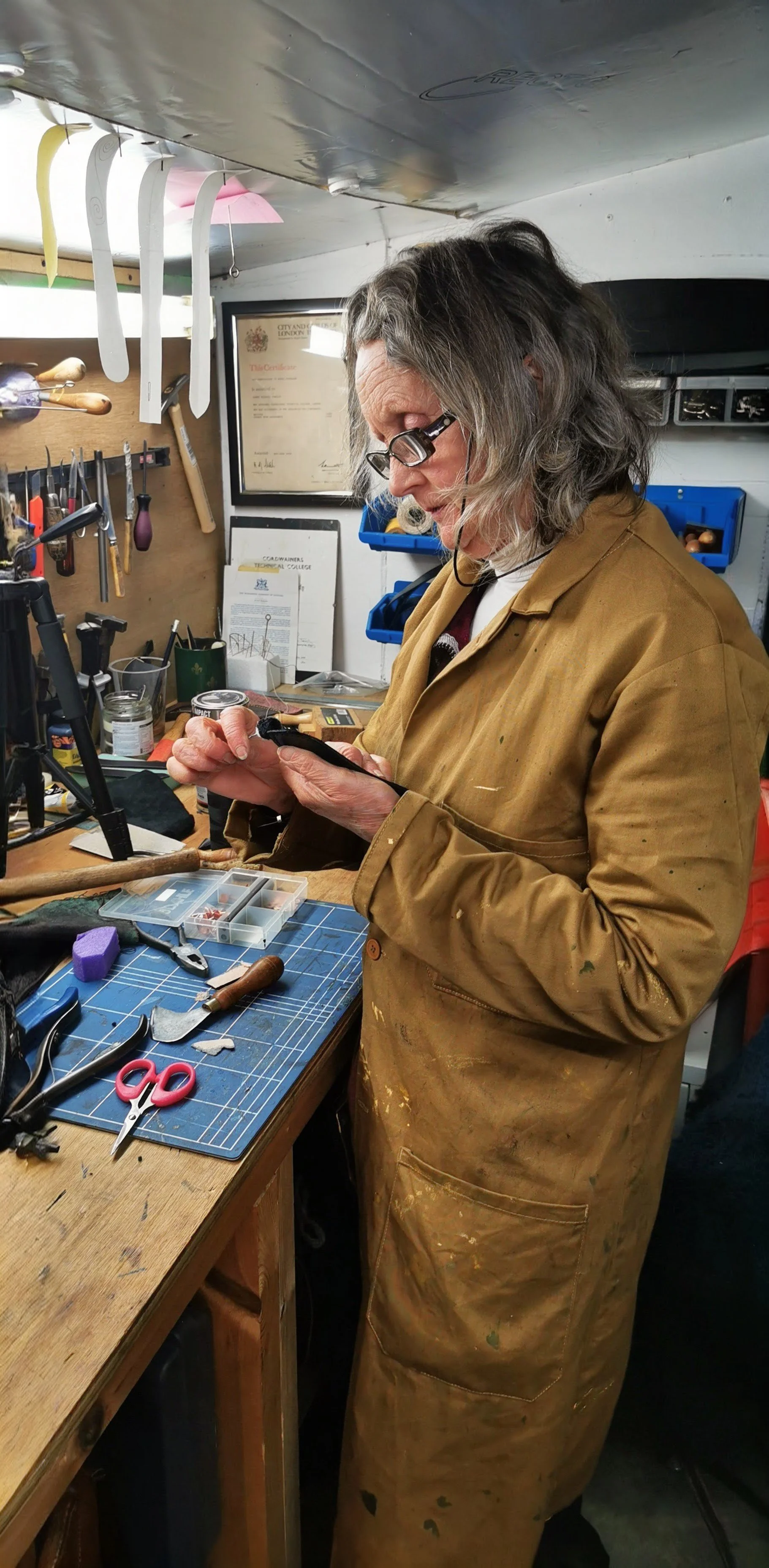 A woman with gray hair and glasses wearing a stained brown work coat, working on a craft project at a cluttered workshop table with tools, scissors, and small containers.