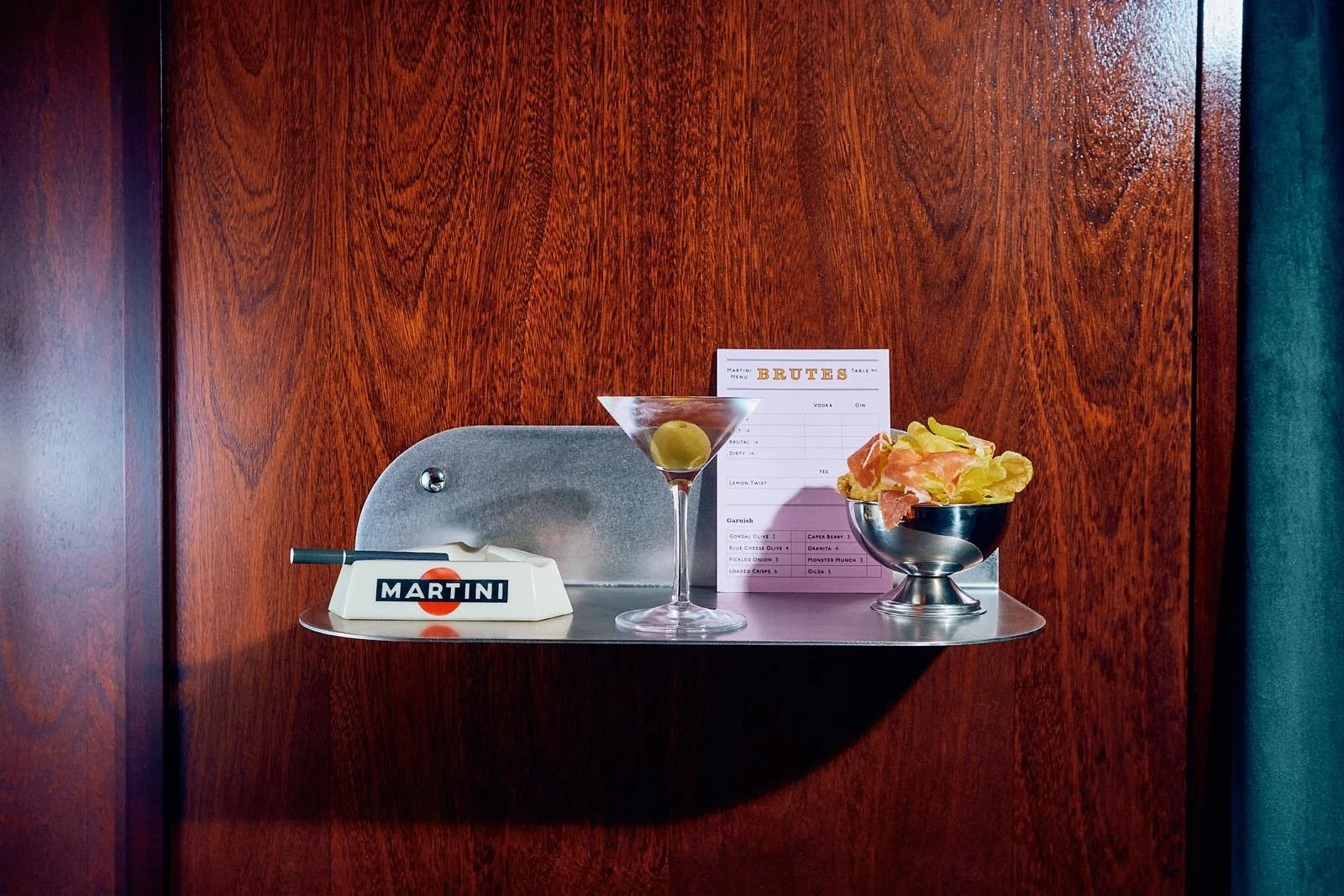 A metallic shelf attached to a wooden wall displays a martini glass with a green olive, a bowl of potato chips, a table order sheet, a white bottle opener with the word 'MARTINI' on it, and a metal chair behind the shelf.