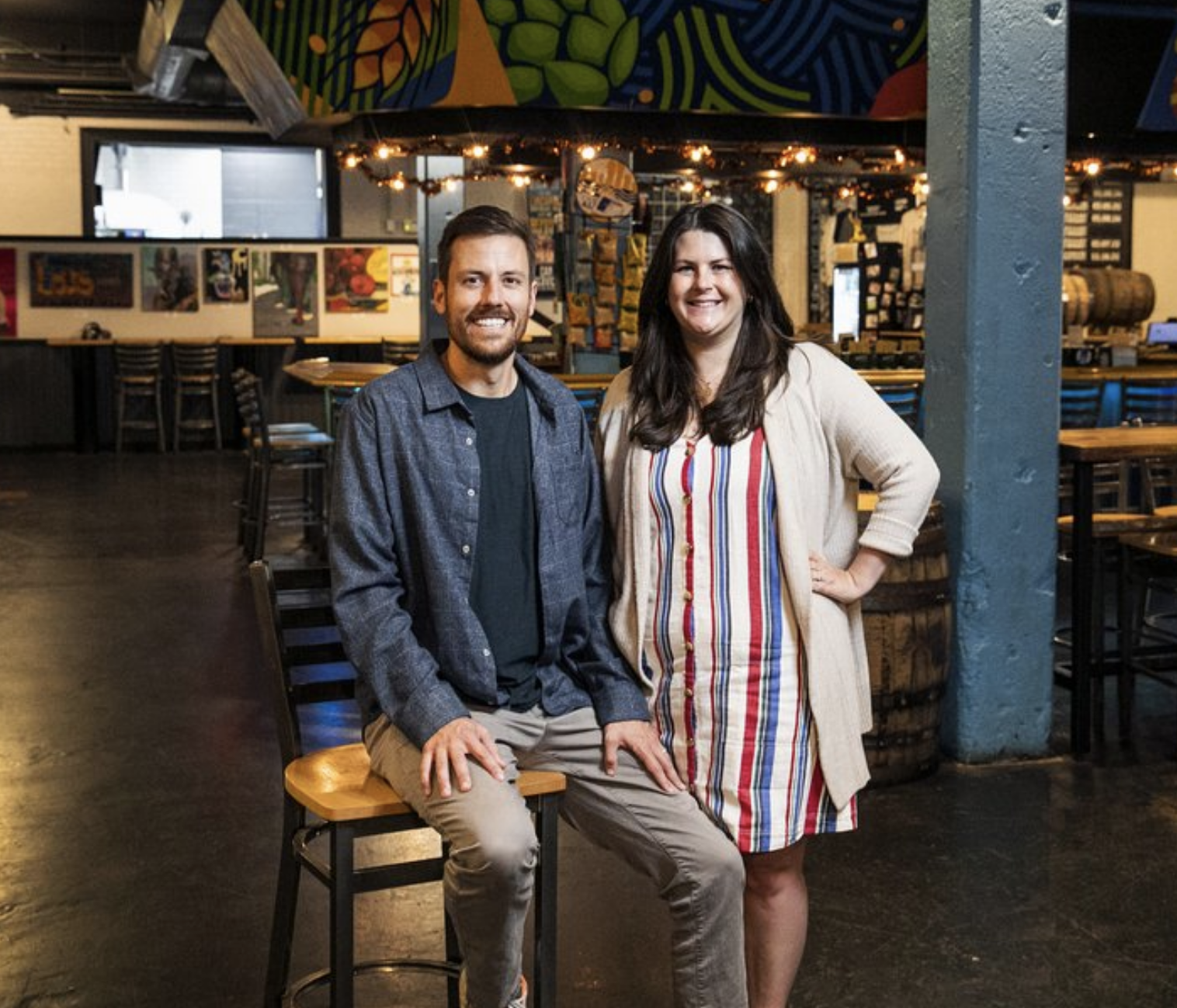 A man and a woman smiling in a bar or restaurant with artwork and barrels in the background.