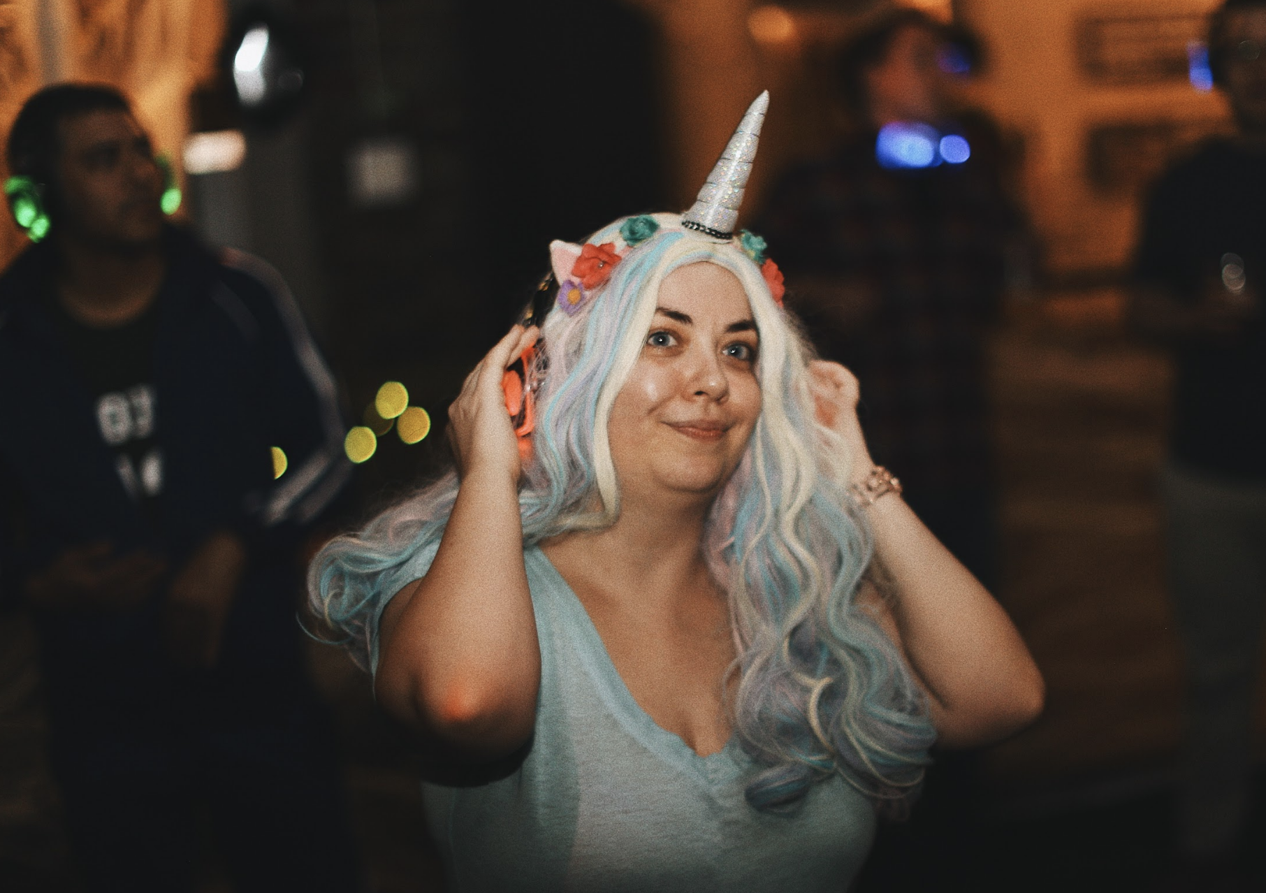 A woman wearing a unicorn-themed costume, including a unicorn horn headband with rainbow-colored hair, in a party setting with blurred background.
