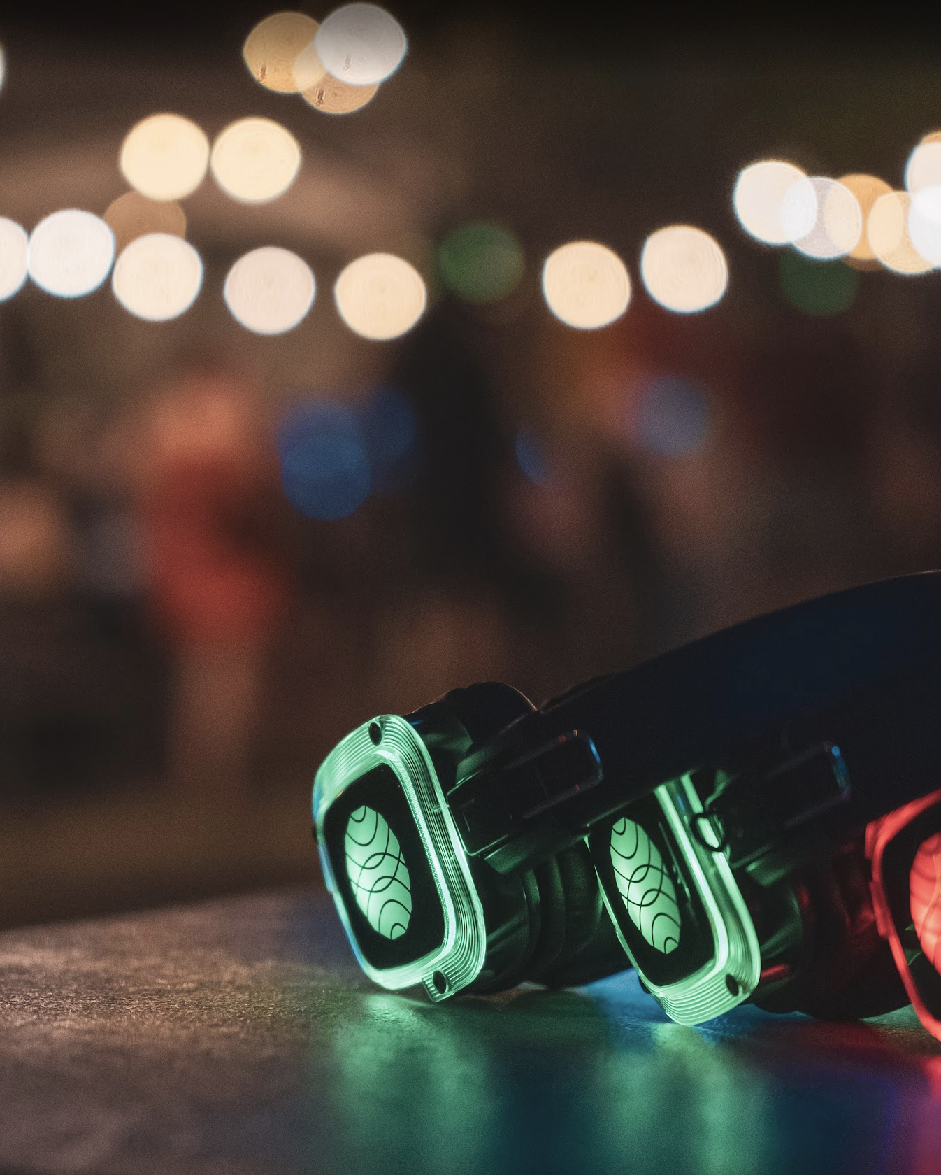 Close-up of gaming glasses with illuminated green and red frames, resting on a surface at night with blurred city lights in the background.