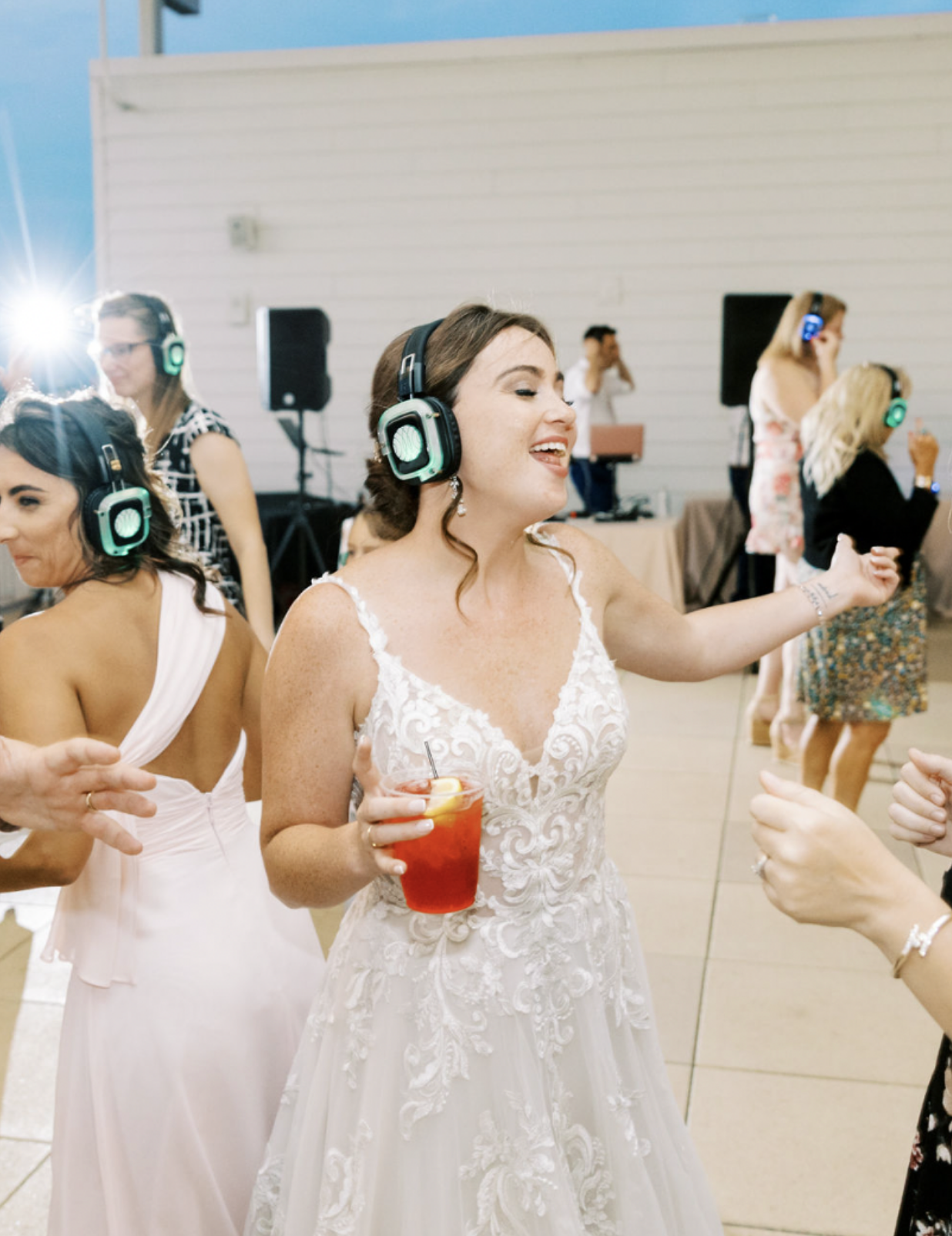 Bride in a white lace wedding dress dancing at her wedding reception, wearing headphones, holding a red drink with lemon, surrounded by guests dancing and enjoying music.