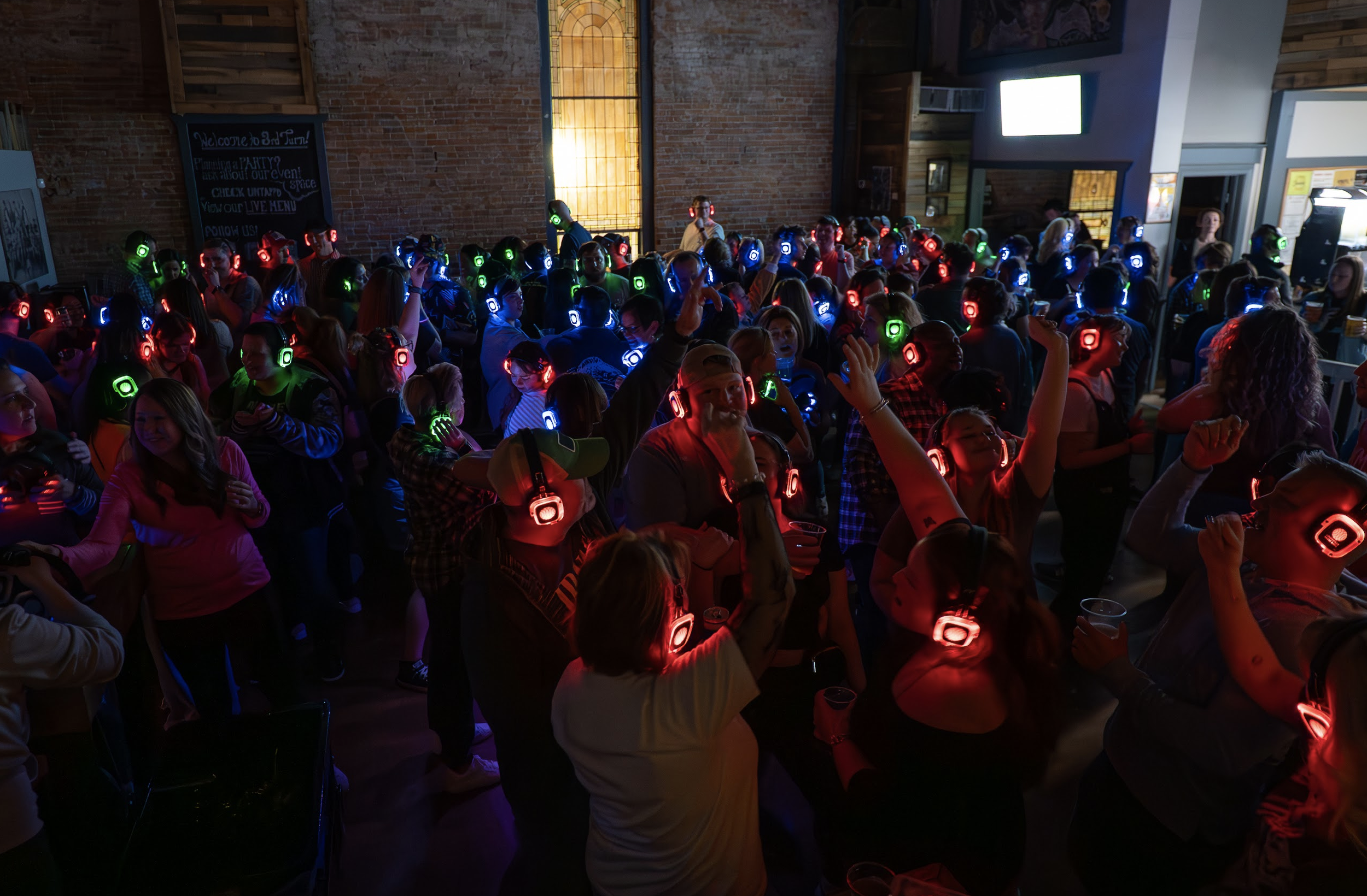 A large group of people in a dimly lit venue, all wearing colorful LED headphones, enjoying a silent party with some people dancing and others holding drinks.