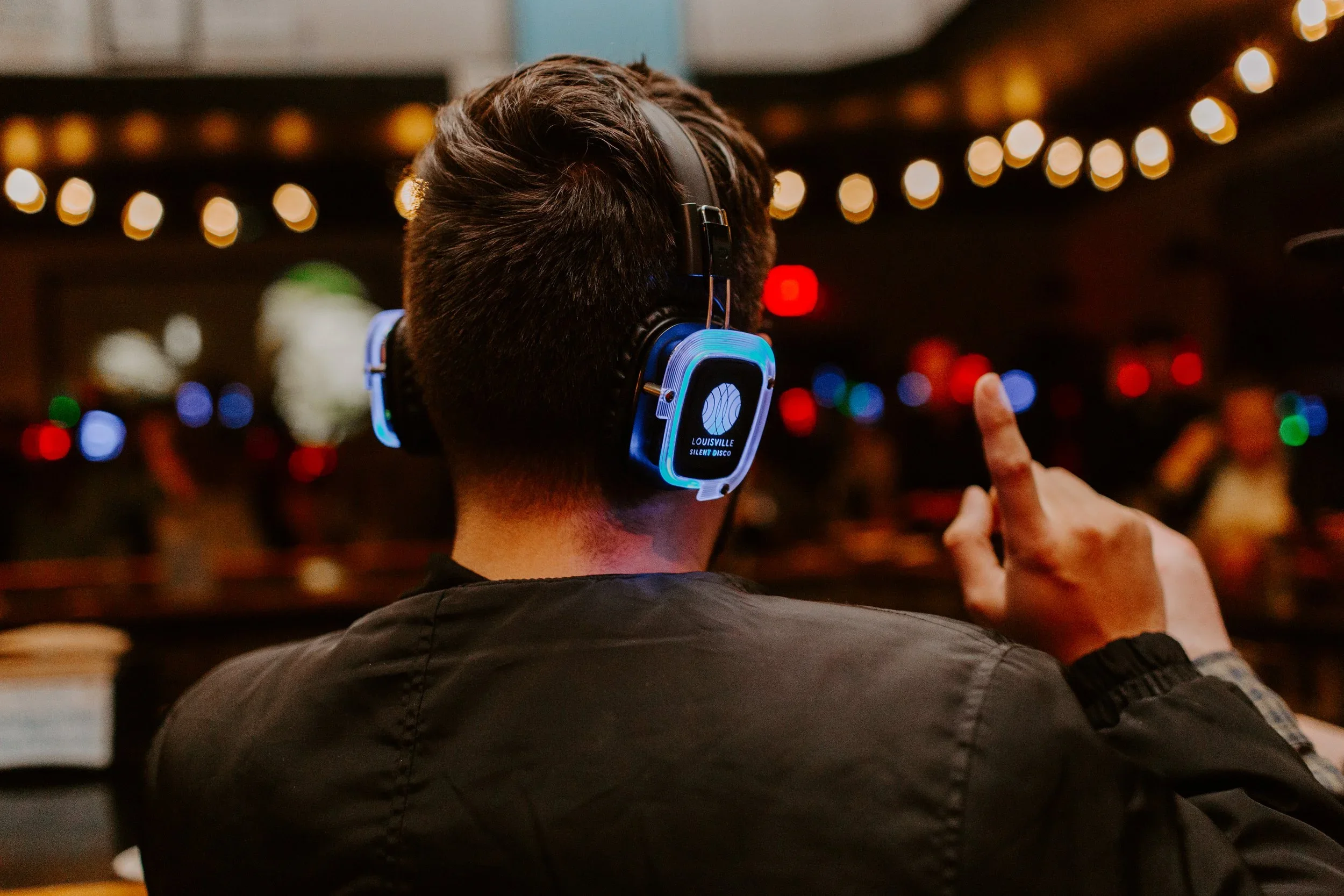 Person wearing headphones at a bar or nightclub, with colorful blurred lights in the background, giving a thumbs-up gesture.
