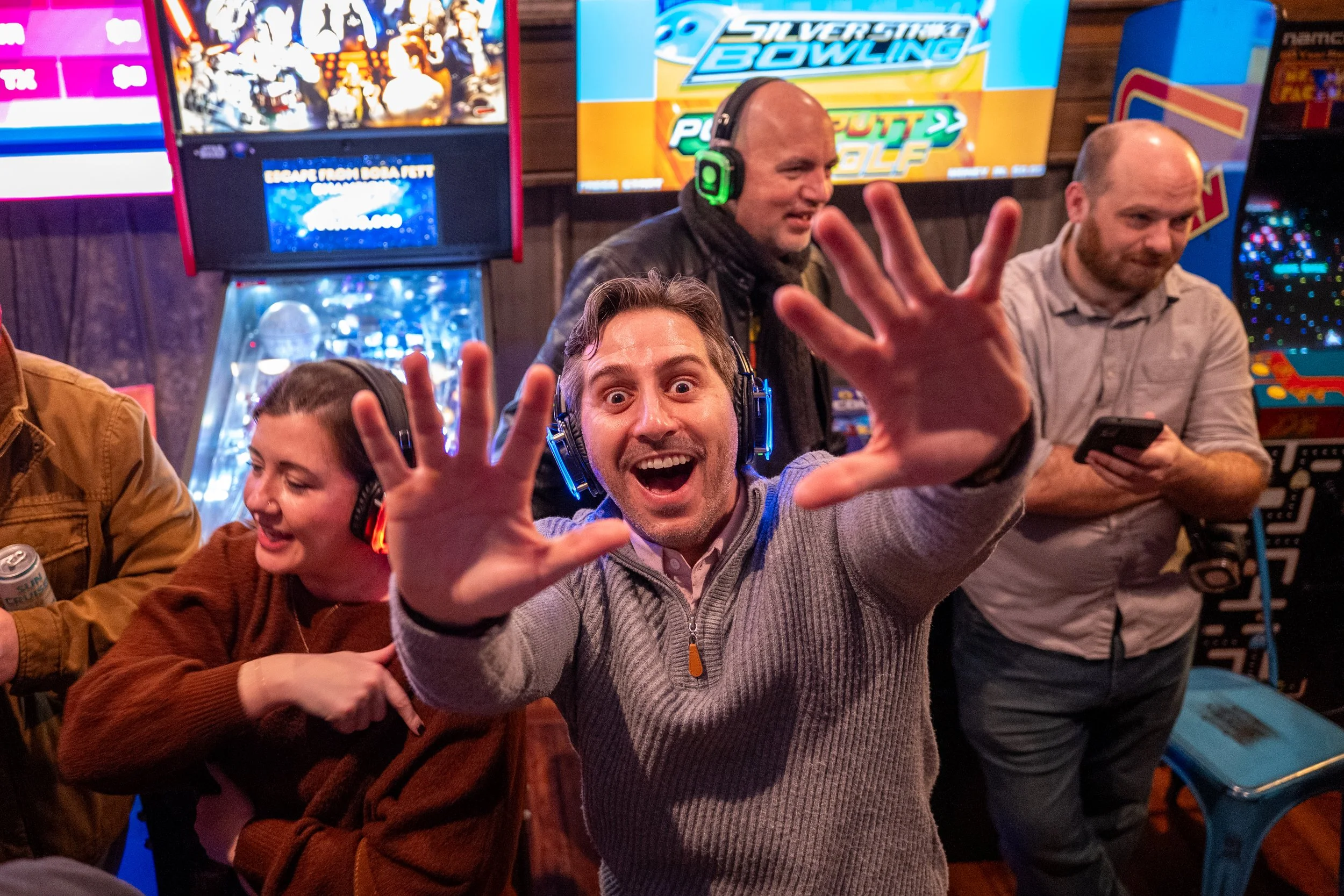 Group of people enjoying arcade games at an arcade, with one man smiling and reaching towards the camera, wearing a gray sweater and headphones, other people wearing headphones and using smartphones, surrounded by arcade machines and colorful screens.
