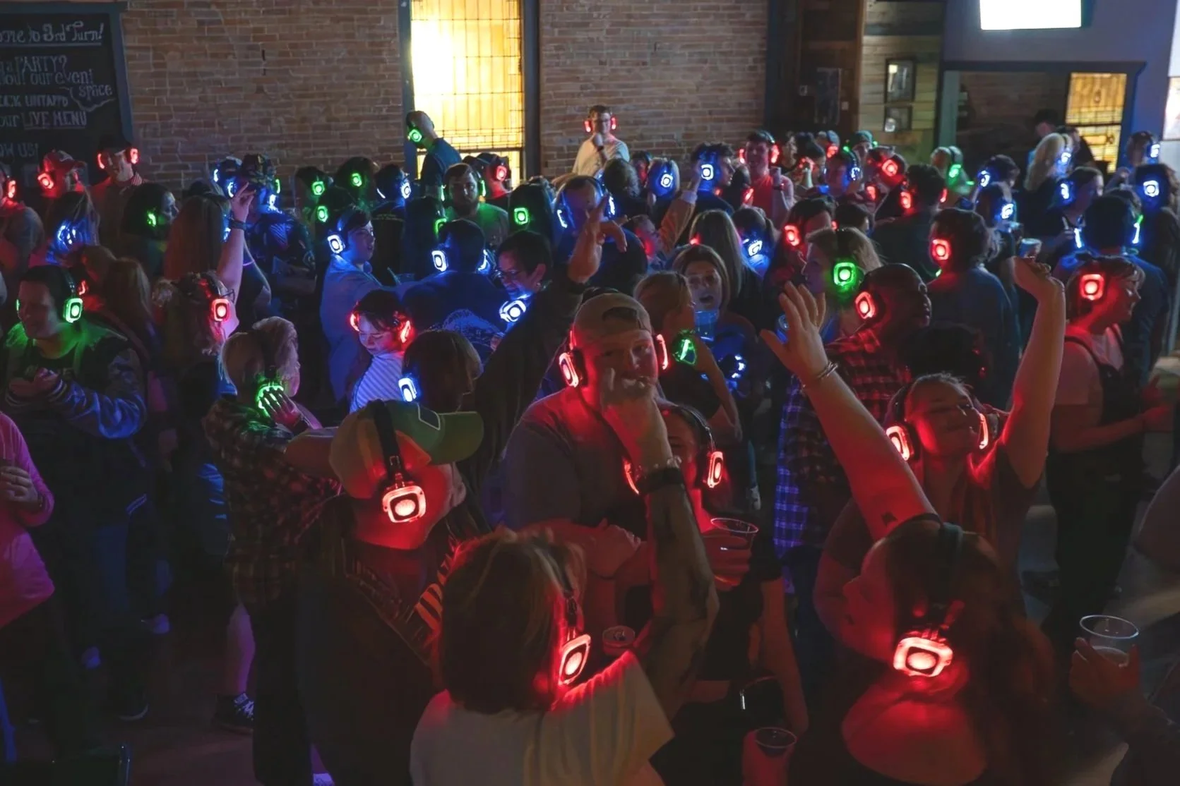 A large group of people wearing colorful glowing headphones dancing and socializing at a party or event in a dimly lit indoor space.
