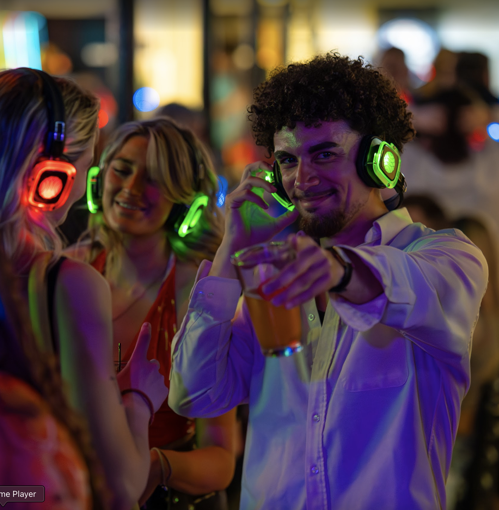 A group of young people at a party wearing LED-lit headphones, with a man in the foreground smiling and pointing at the camera, holding a drink, and a woman smiling in the background.