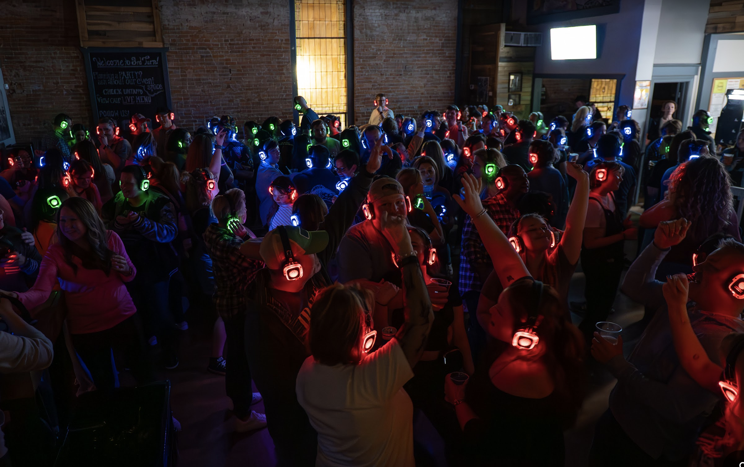 Crowd of people wearing illuminated headphones at a party or event in a dimly lit venue with exposed brick walls.