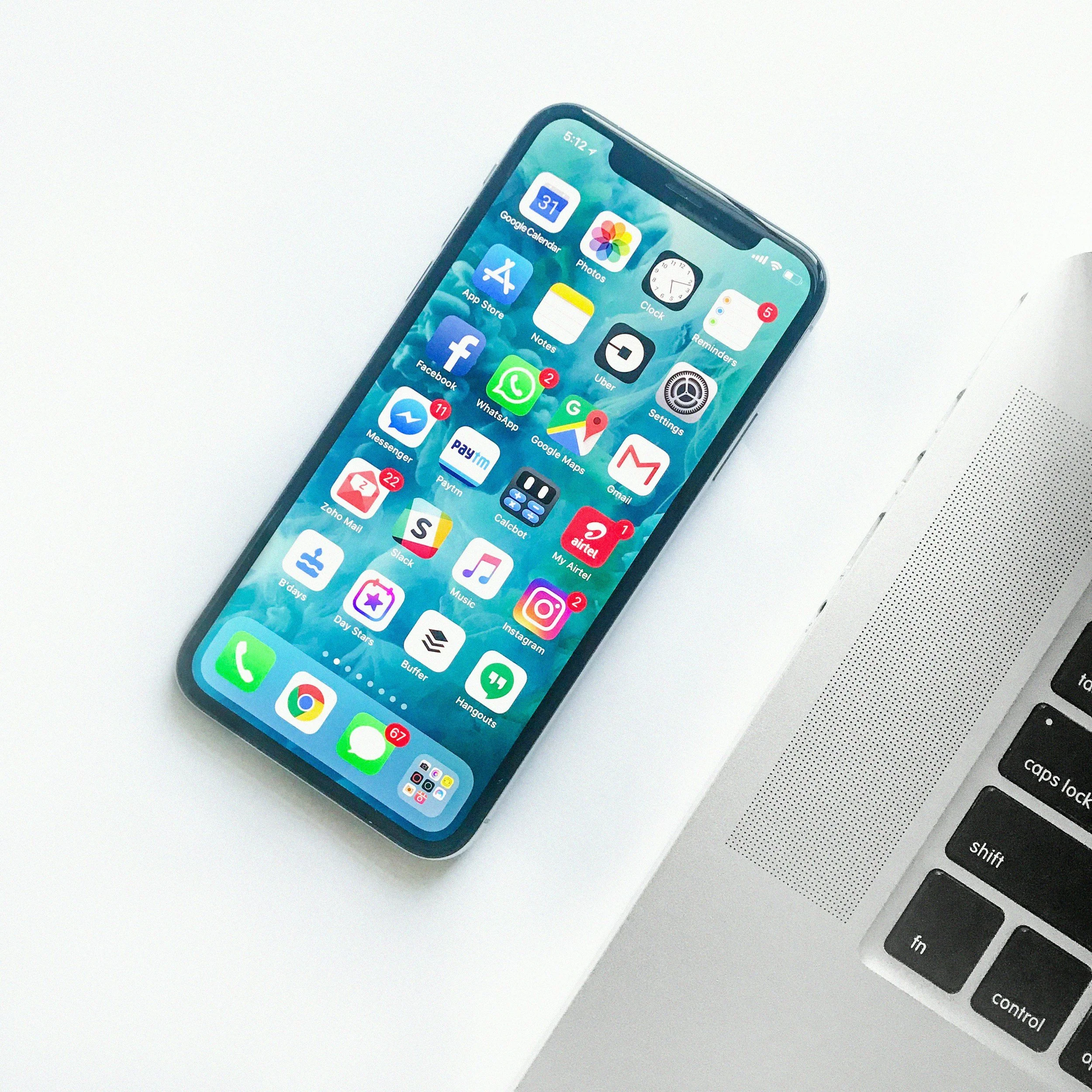An iPhone with various app icons on the screen placed on a white surface next to a silver laptop keyboard.