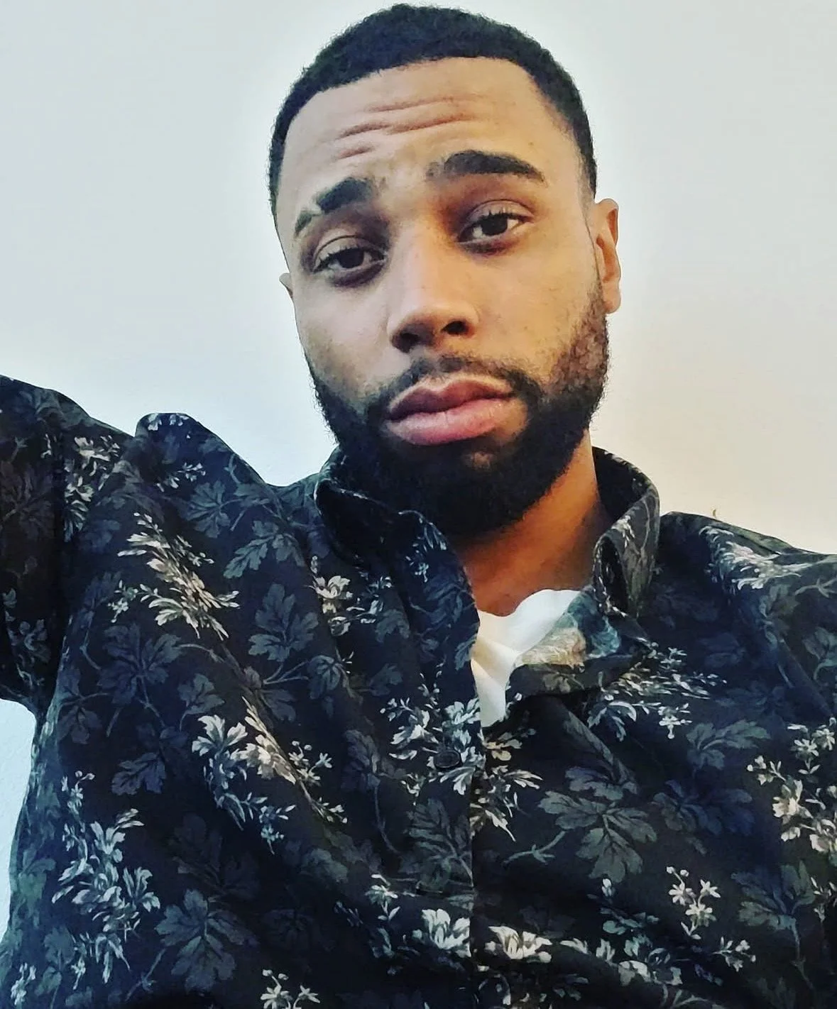 A young man with short curly hair, a beard, and a mustache taking a selfie against a plain light-colored background. He's wearing a dark blue shirt with a floral pattern and a white shirt underneath.