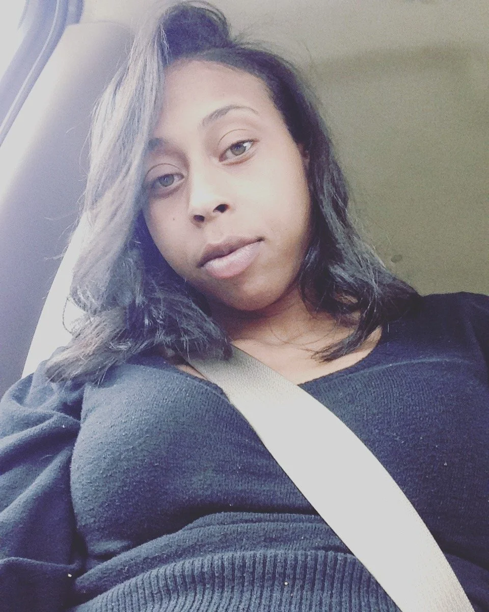 A young woman with black hair and light brown eyes wearing a black shirt and a beige seatbelt, sitting in a car.