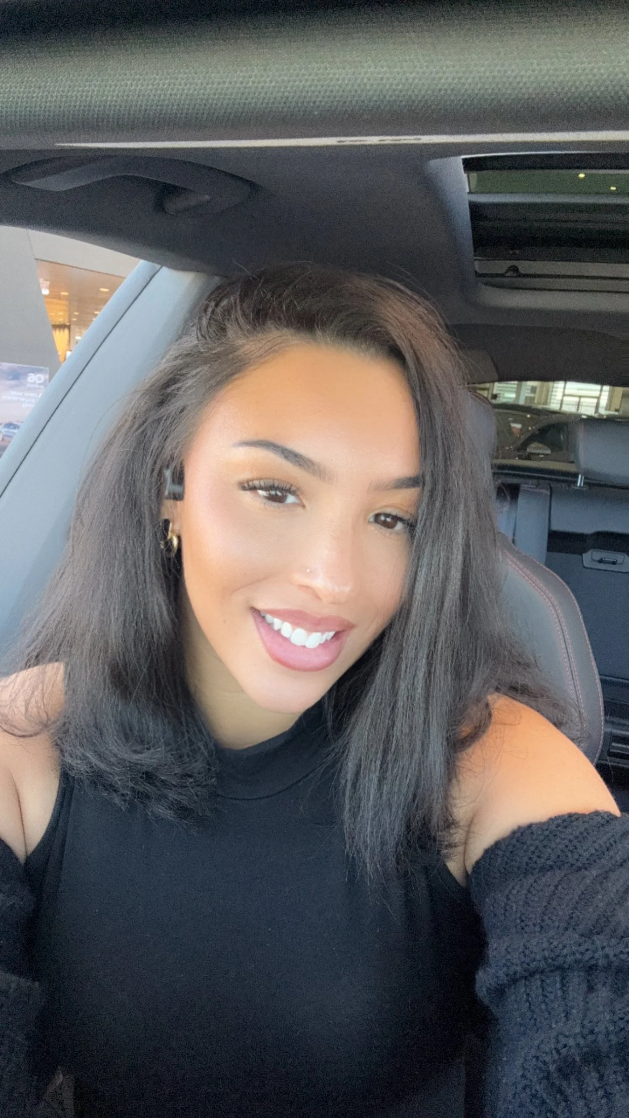 A woman with long dark hair, smiling, sitting inside a car.